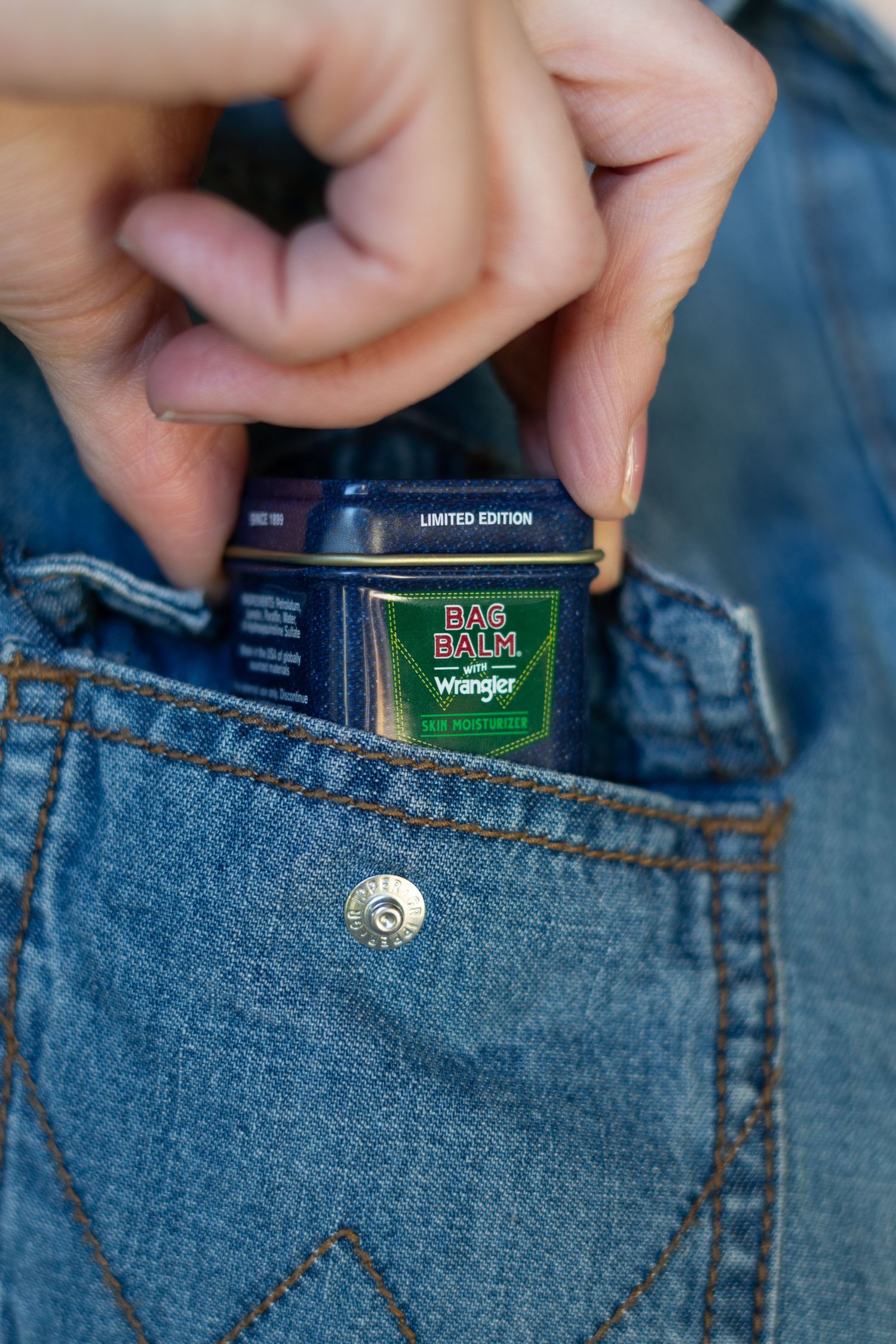 Hand placing limited edition Bag Balm x Wrangler moisturizer tin into denim jeans front pocket.