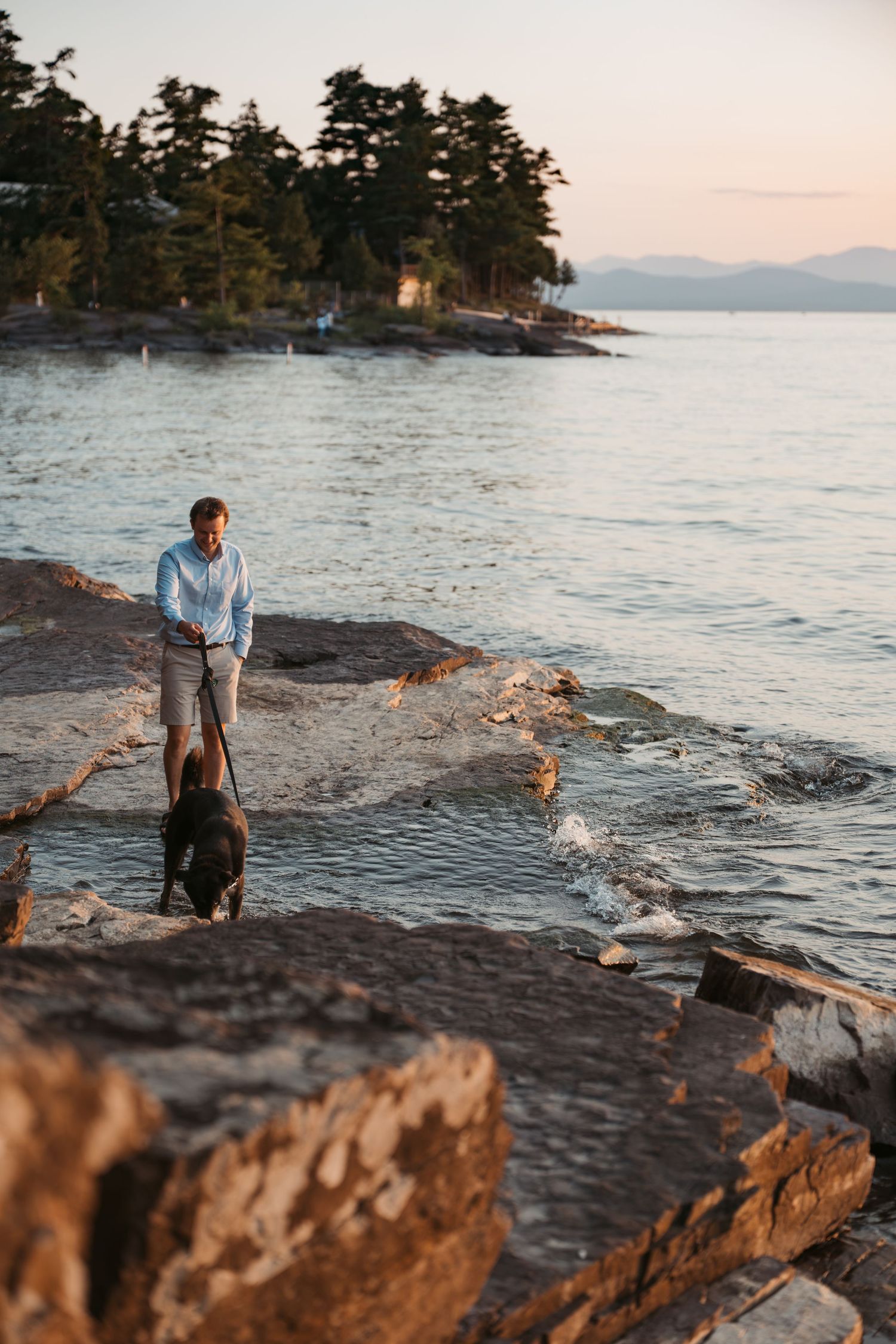 Man walking a dark dog on rocky shoreline at sunset with forested island and mountains in background.