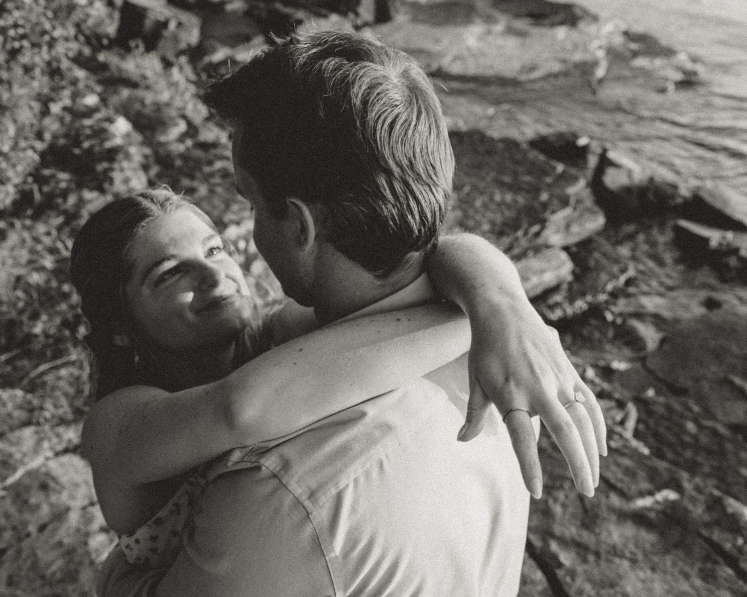 Black and white photo of smiling woman hugging man on rocky shoreline.