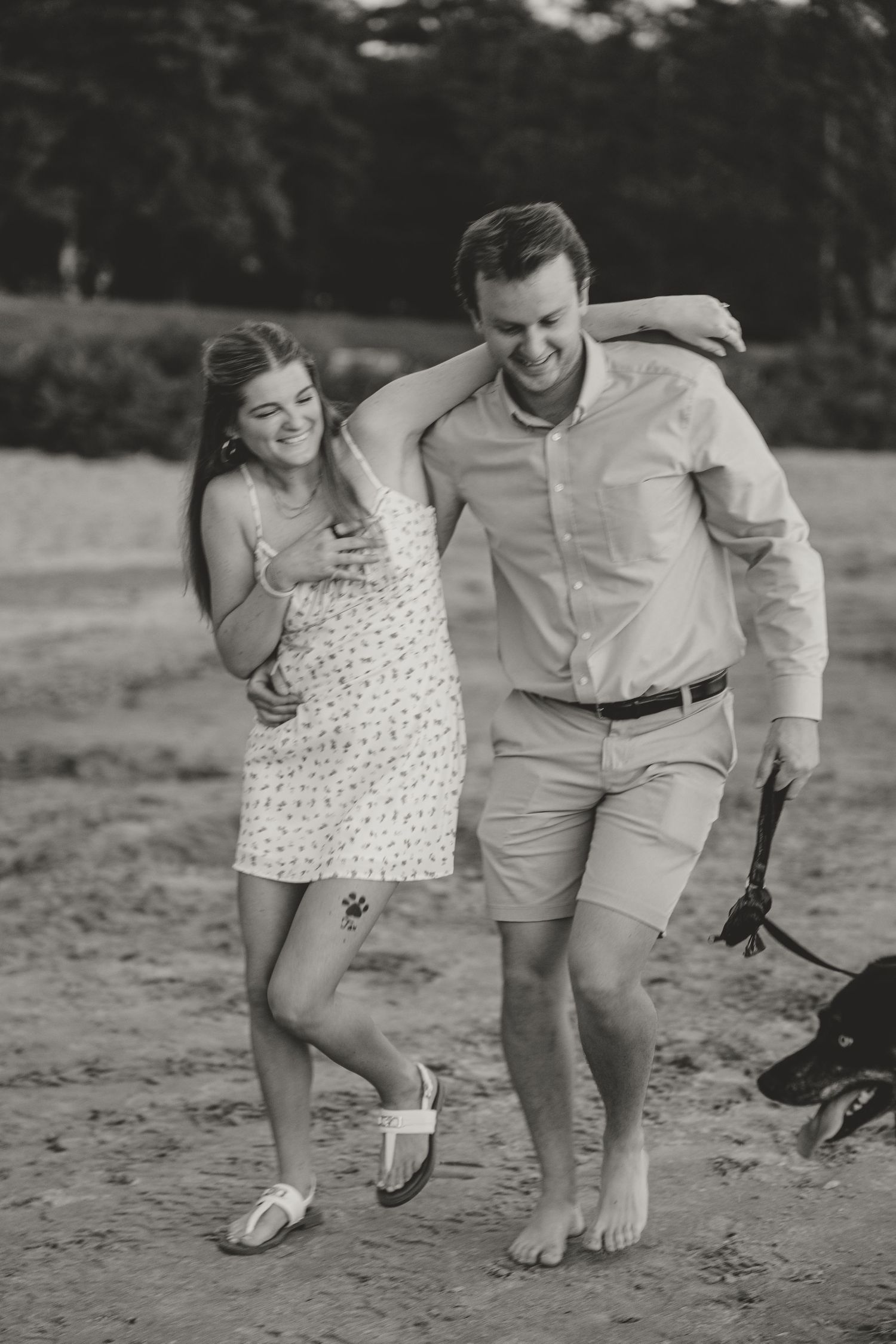Black and white photo of a laughing couple walking arm in arm on a beach.