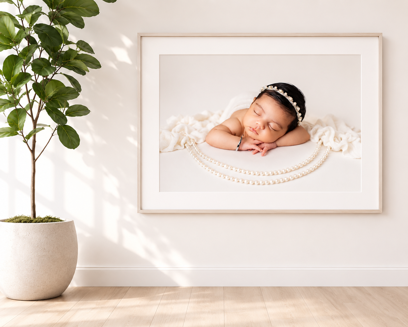 Framed newborn portrait of a sleeping baby with pearl accessories, displayed on a white wall beside a green potted plant.