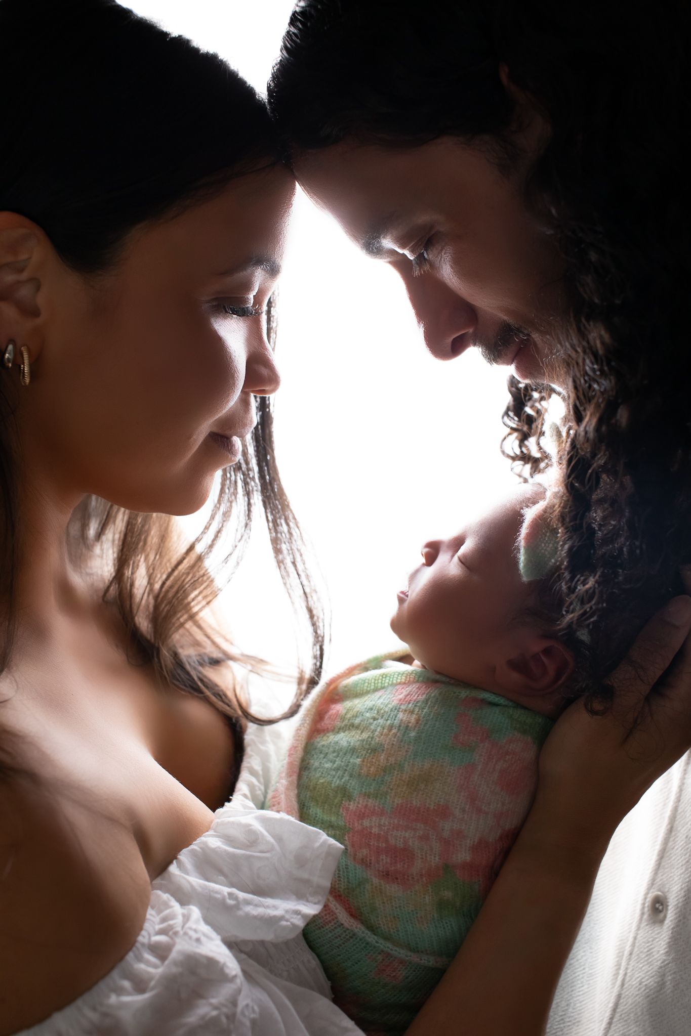 Silhouetted parents lovingly gazing at their awake newborn baby in a dramatic backlit portrait.