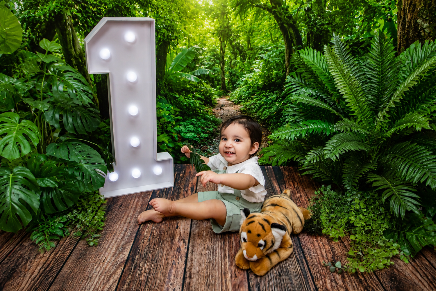 Happy baby boy sitting beside illuminated number one sign with tiger toy in jungle setting.