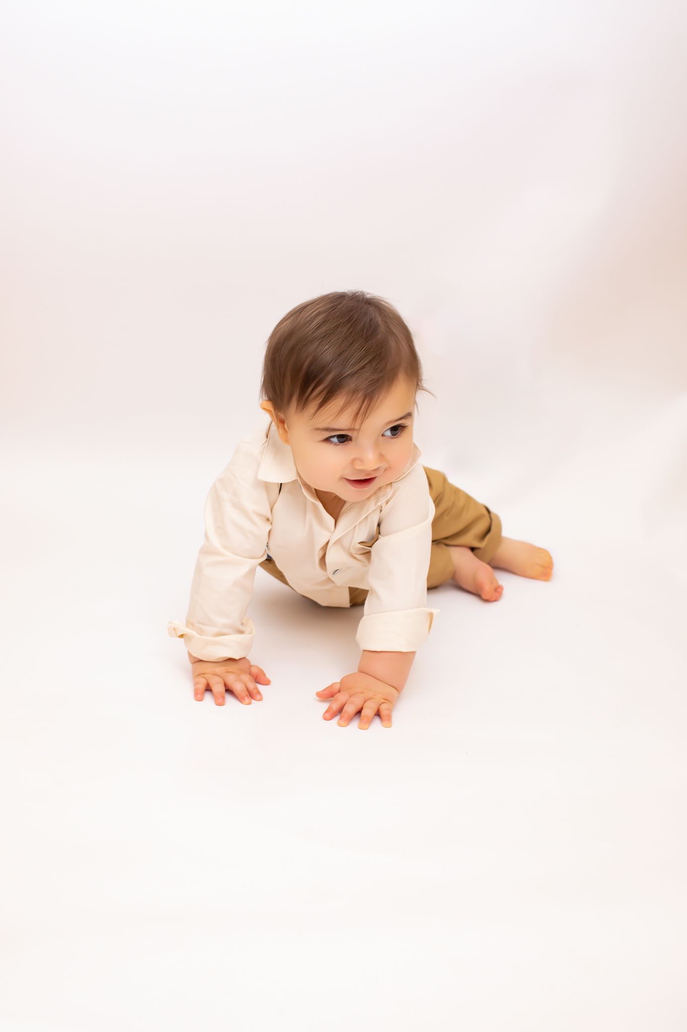 A curious baby in a cream onesie and khaki shorts crawls on a white background.