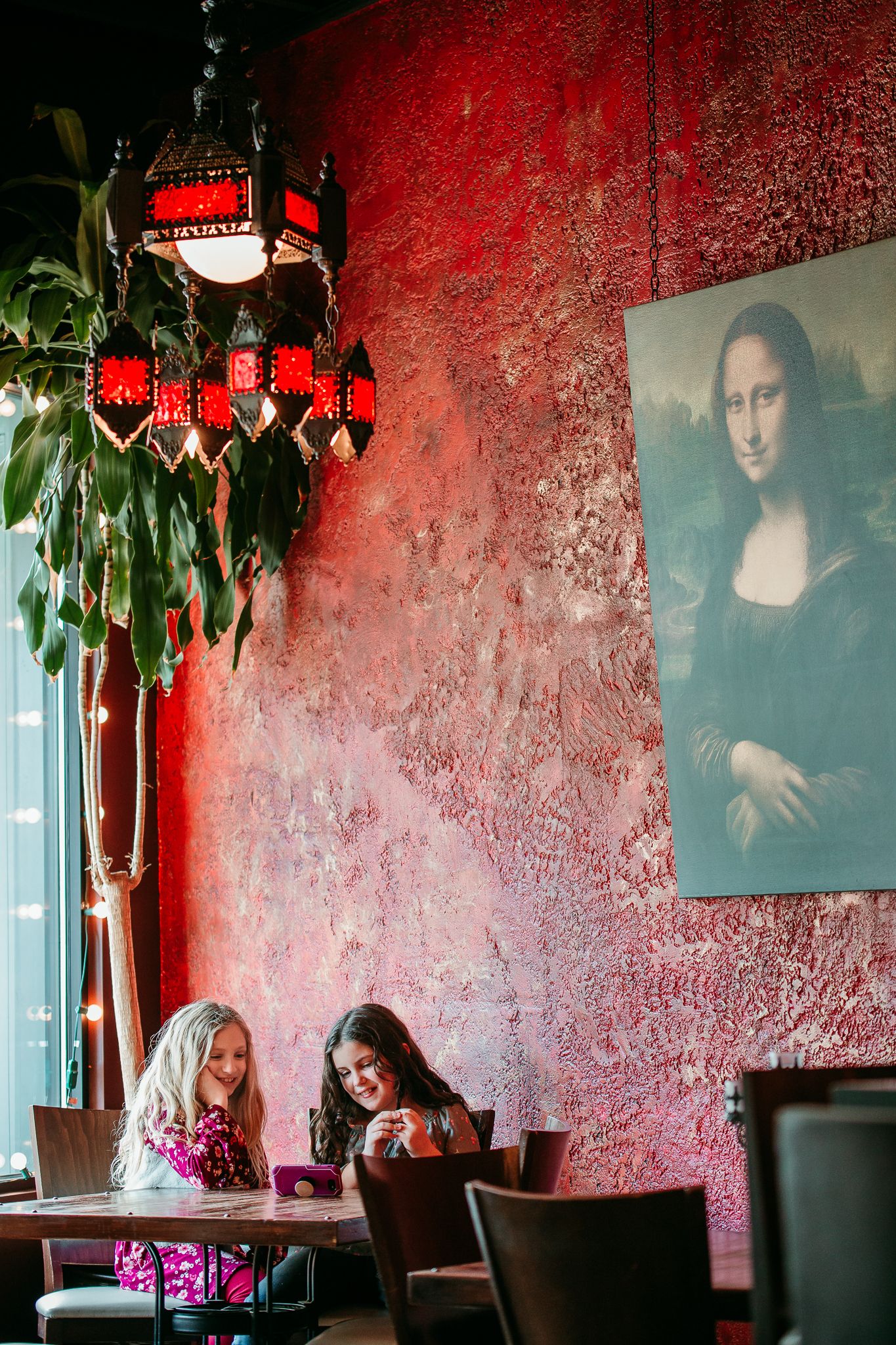 Two young girls sit at a table beneath a Mona Lisa painting in a red-walled restaurant.