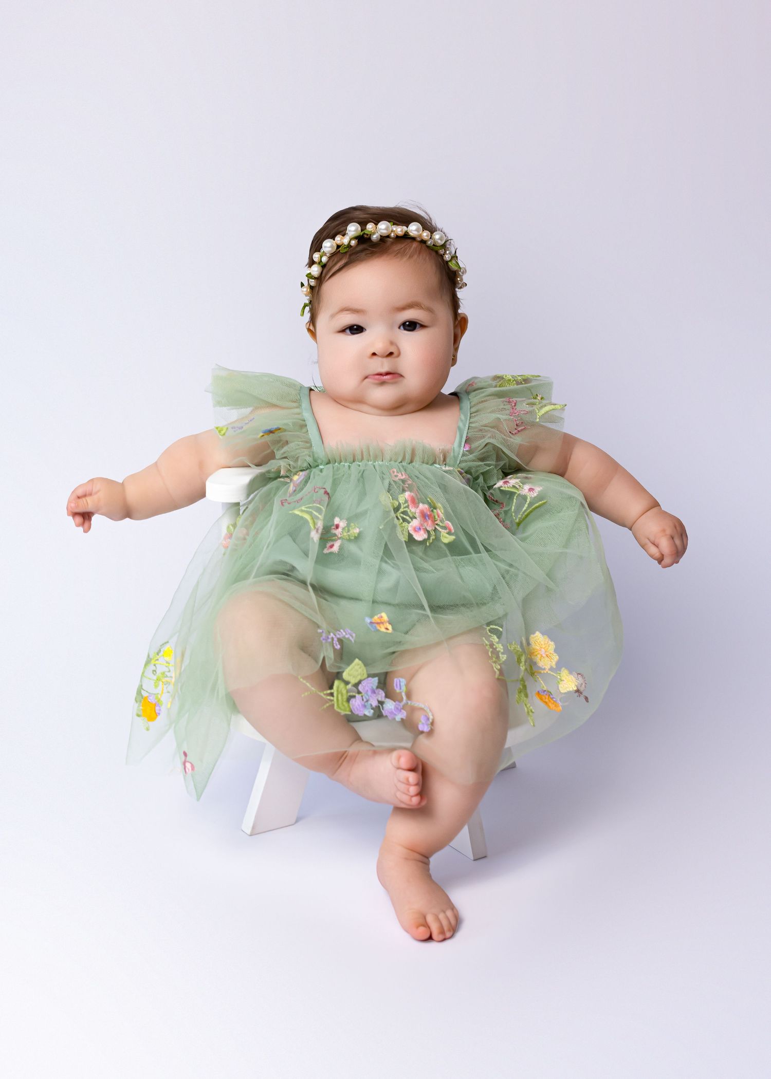 Baby girl in green floral tulle dress and pearl headband sitting on white stool during professional photo session.