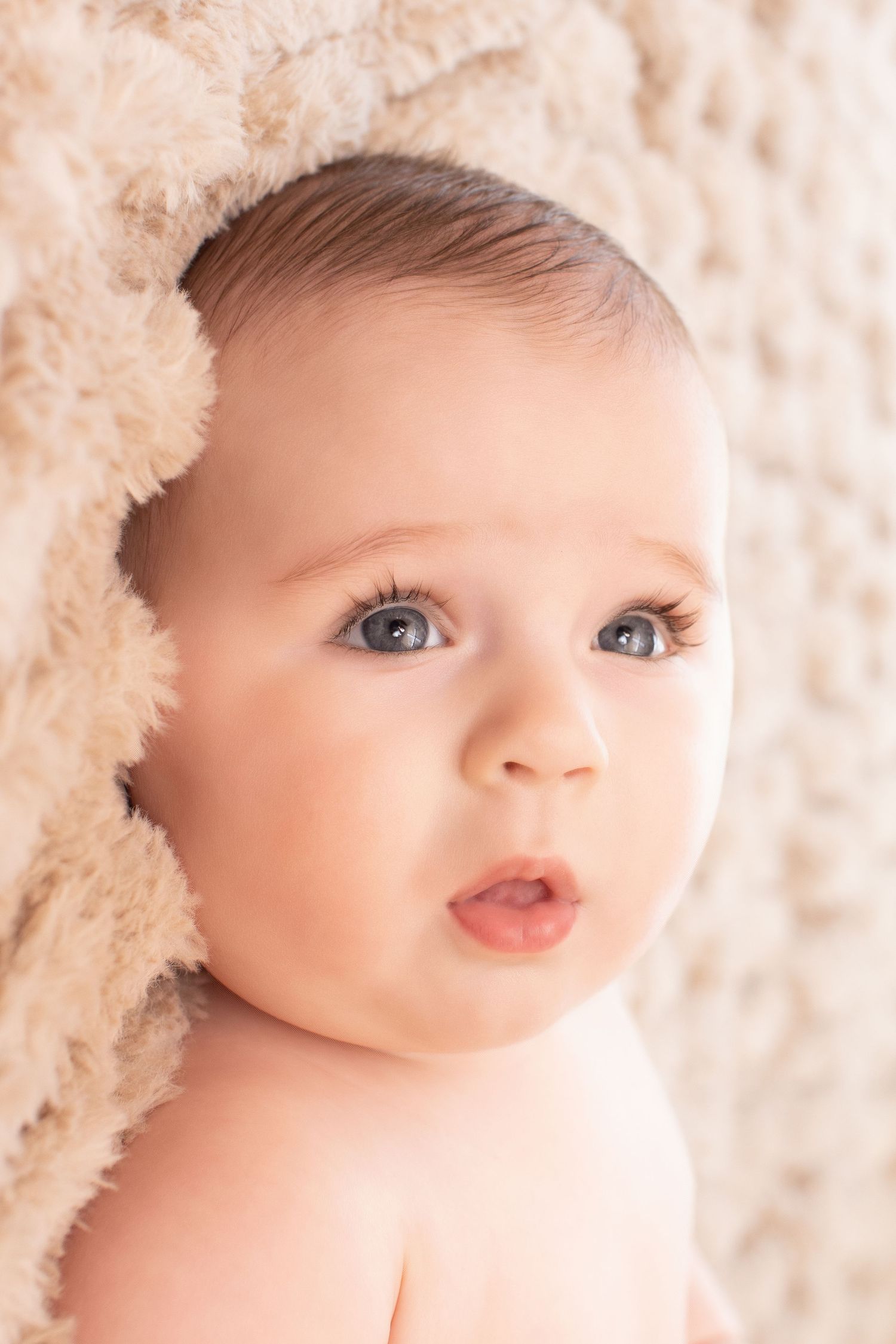 Adorable baby with blue eyes peeks out from under a soft beige blanket with a curious expression.