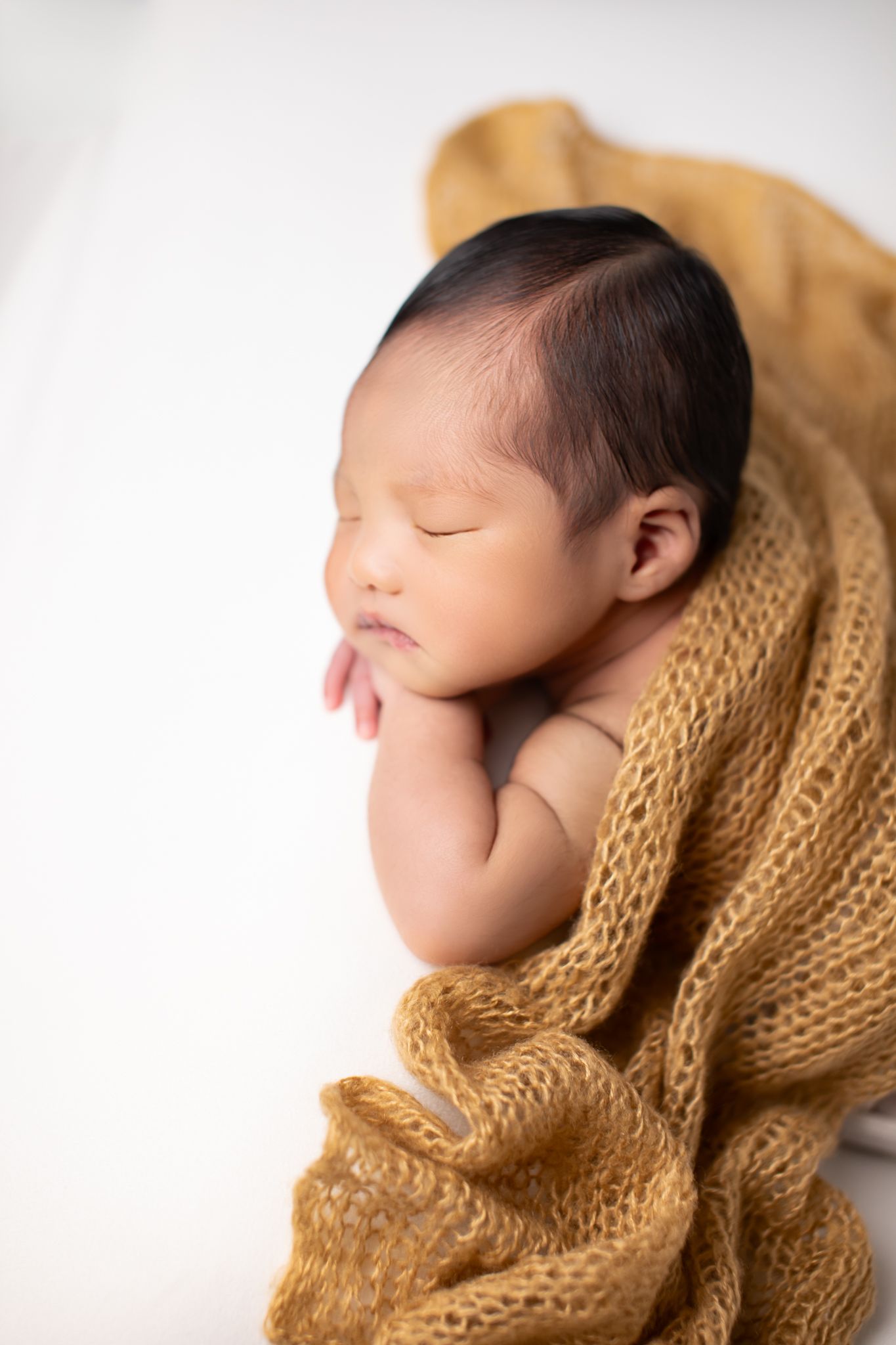 Close-up of sleeping newborn baby wrapped in a golden mustard knit blanket on a white background.