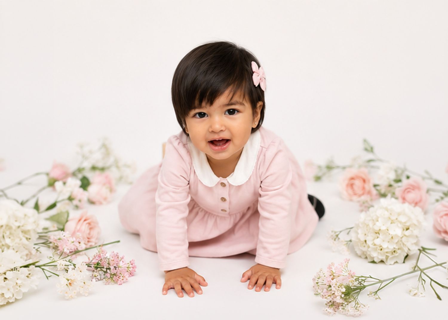 Happy toddler girl in pink dress crawling among pink roses and white flowers on white background.