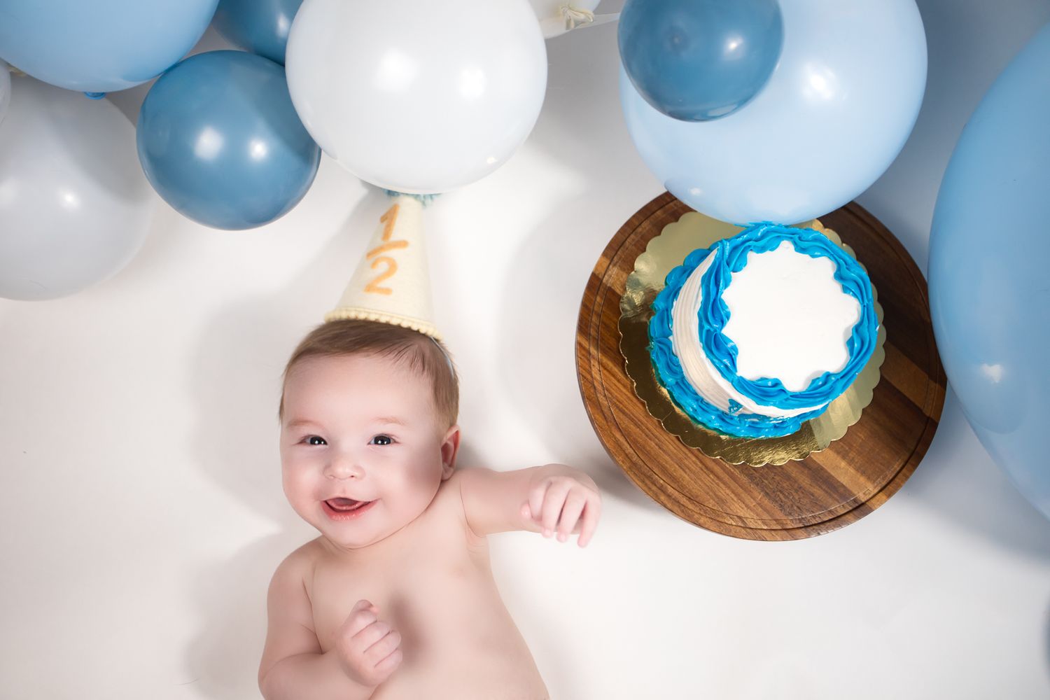 Laughing baby boy in birthday hat lying near blue frosted cake with blue balloons backdrop.