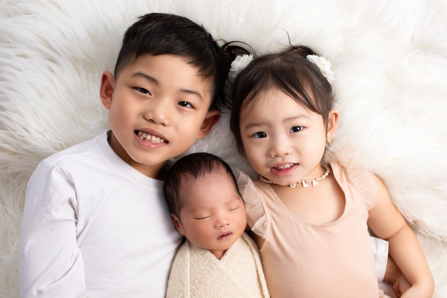 Two smiling siblings lying with a sleeping newborn baby on a white fluffy blanket.