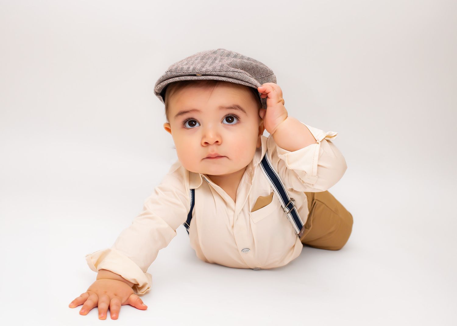 Adorable baby boy in vintage cap and suspenders posing on white studio floor.