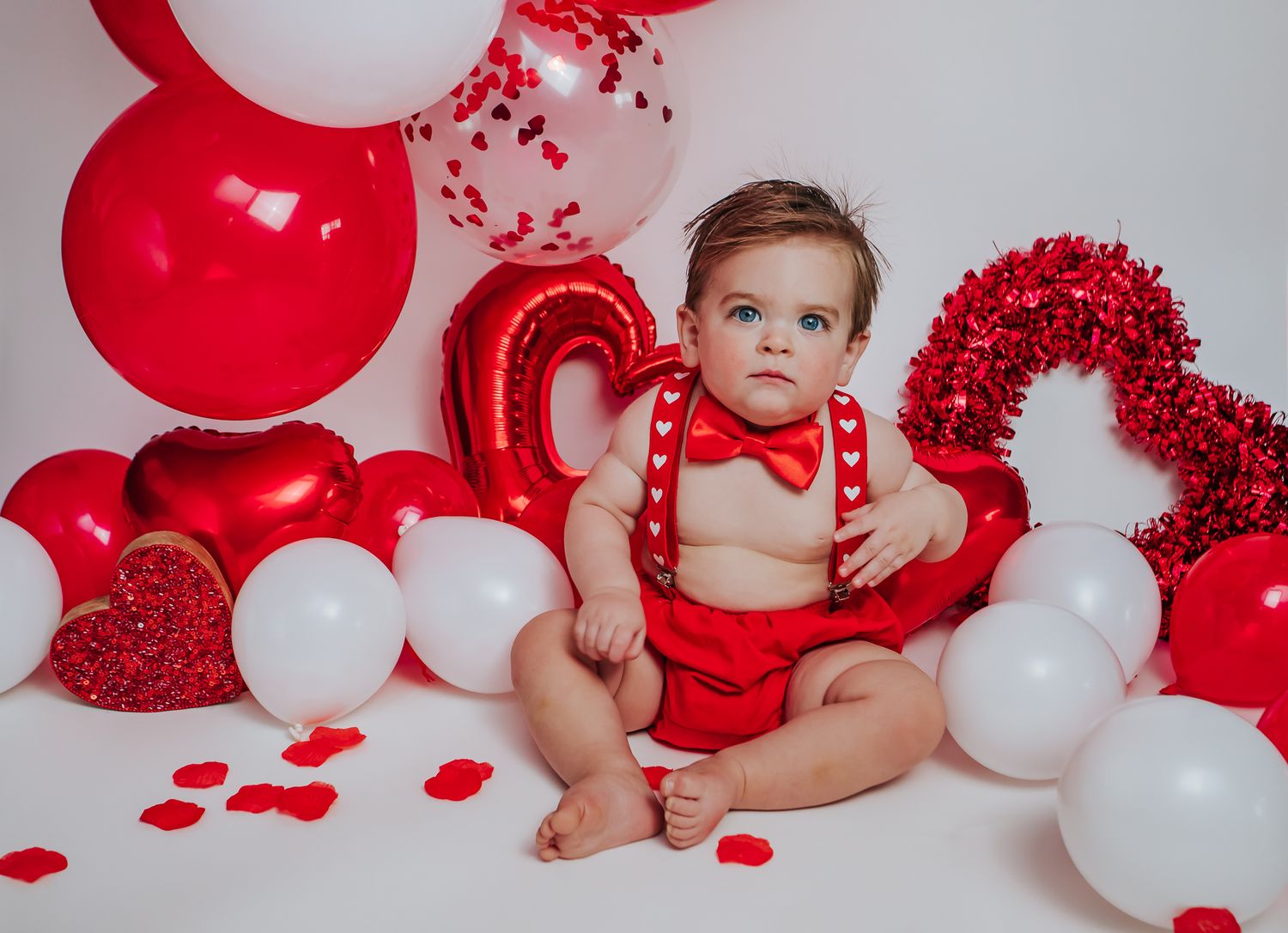 Bright-eyed baby in red suspenders and bow tie posed with red and white balloons and heart decorations.