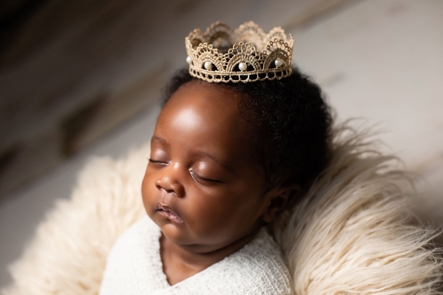 Peaceful newborn baby wearing a gold lace crown resting on a fluffy white blanket.