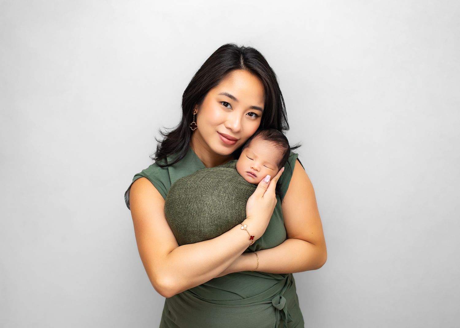 Smiling Asian mother in green dress holds sleeping swaddled newborn baby against her chest.