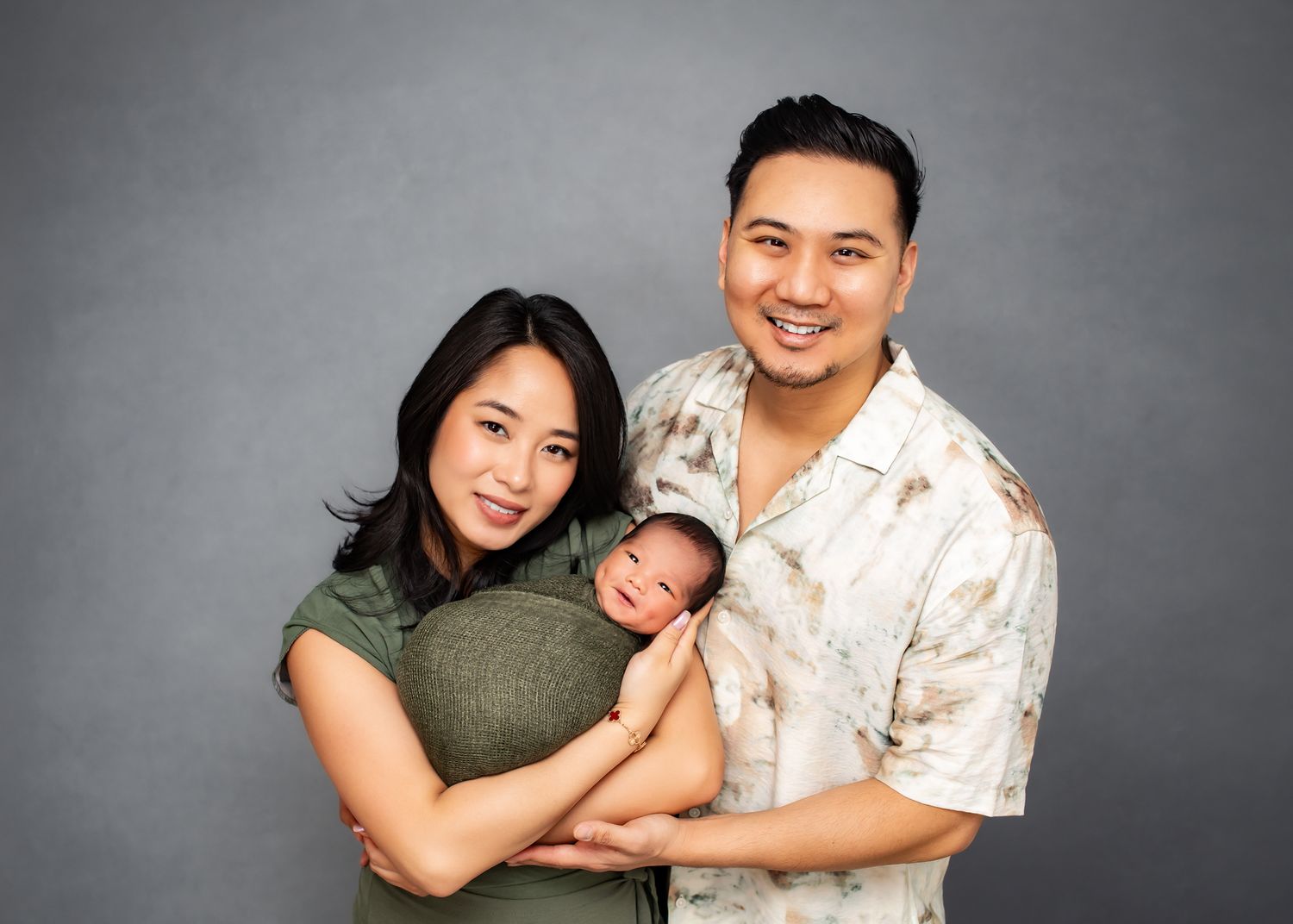 Smiling Asian family portrait with mother and father holding their swaddled newborn baby.