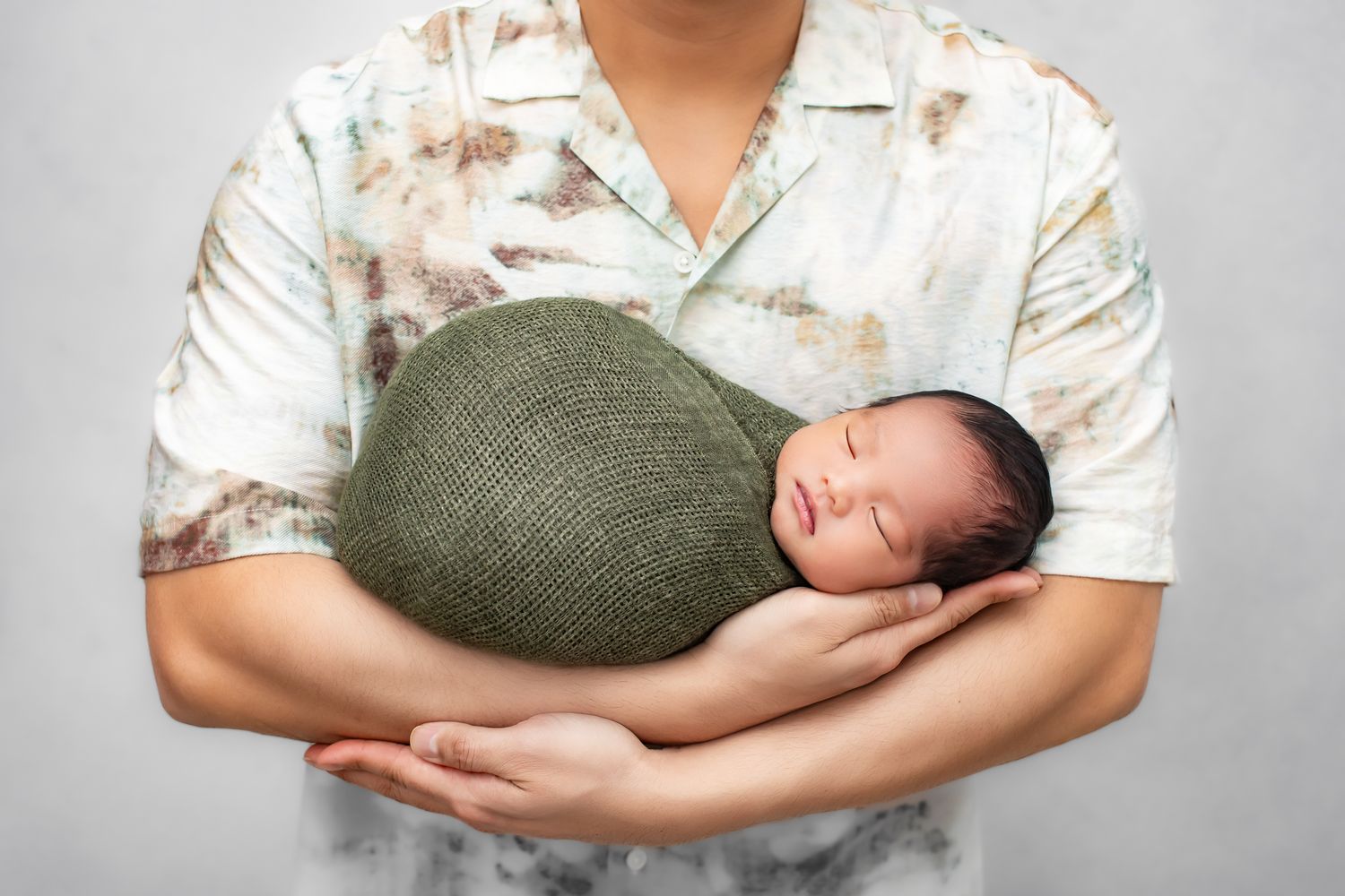 Father in floral shirt gently cradles sleeping newborn wrapped in green knit swaddle in his arms.