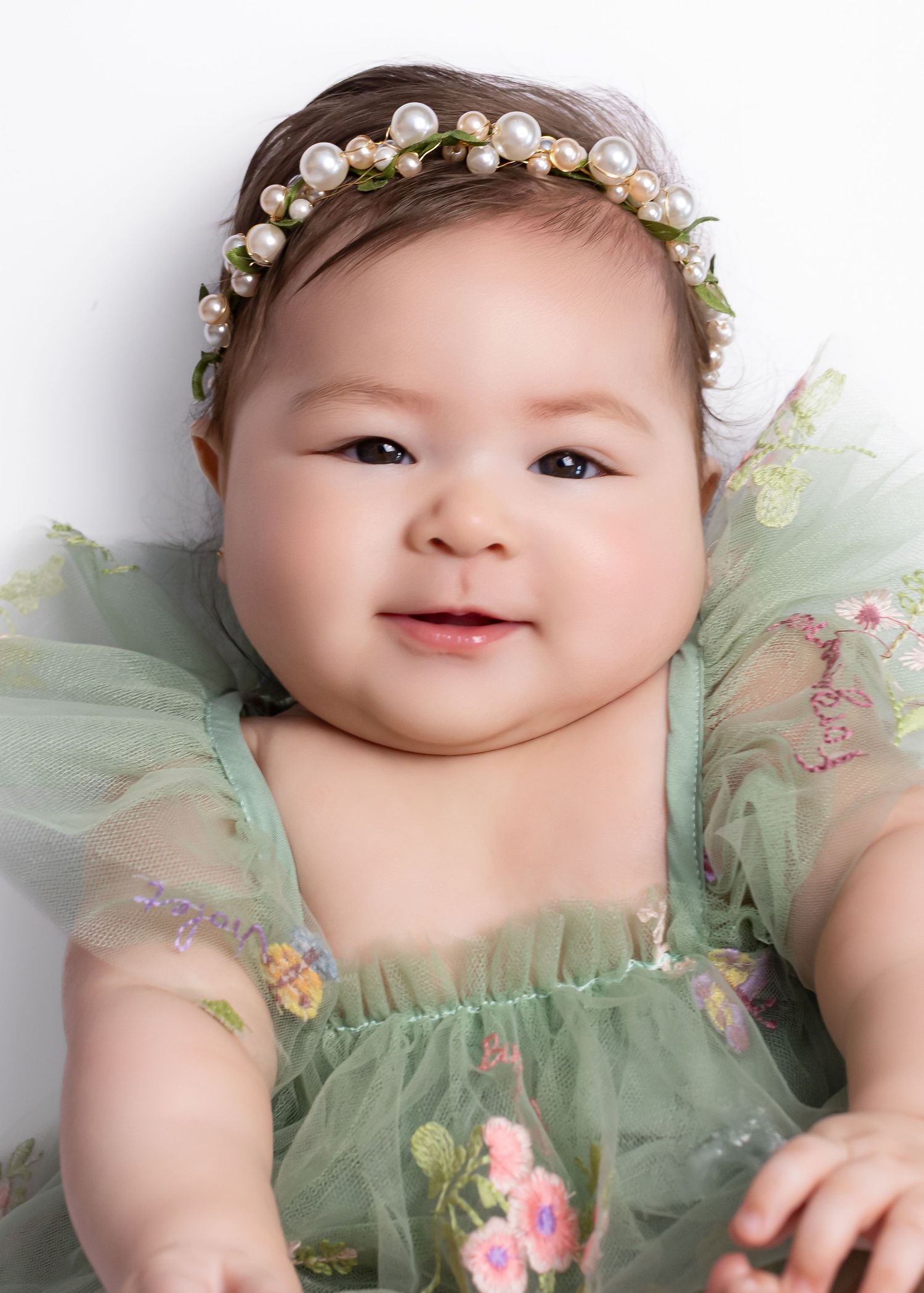 Adorable baby girl in a green floral tulle dress wearing a pearl and flower headband, smiling sweetly at the camera.