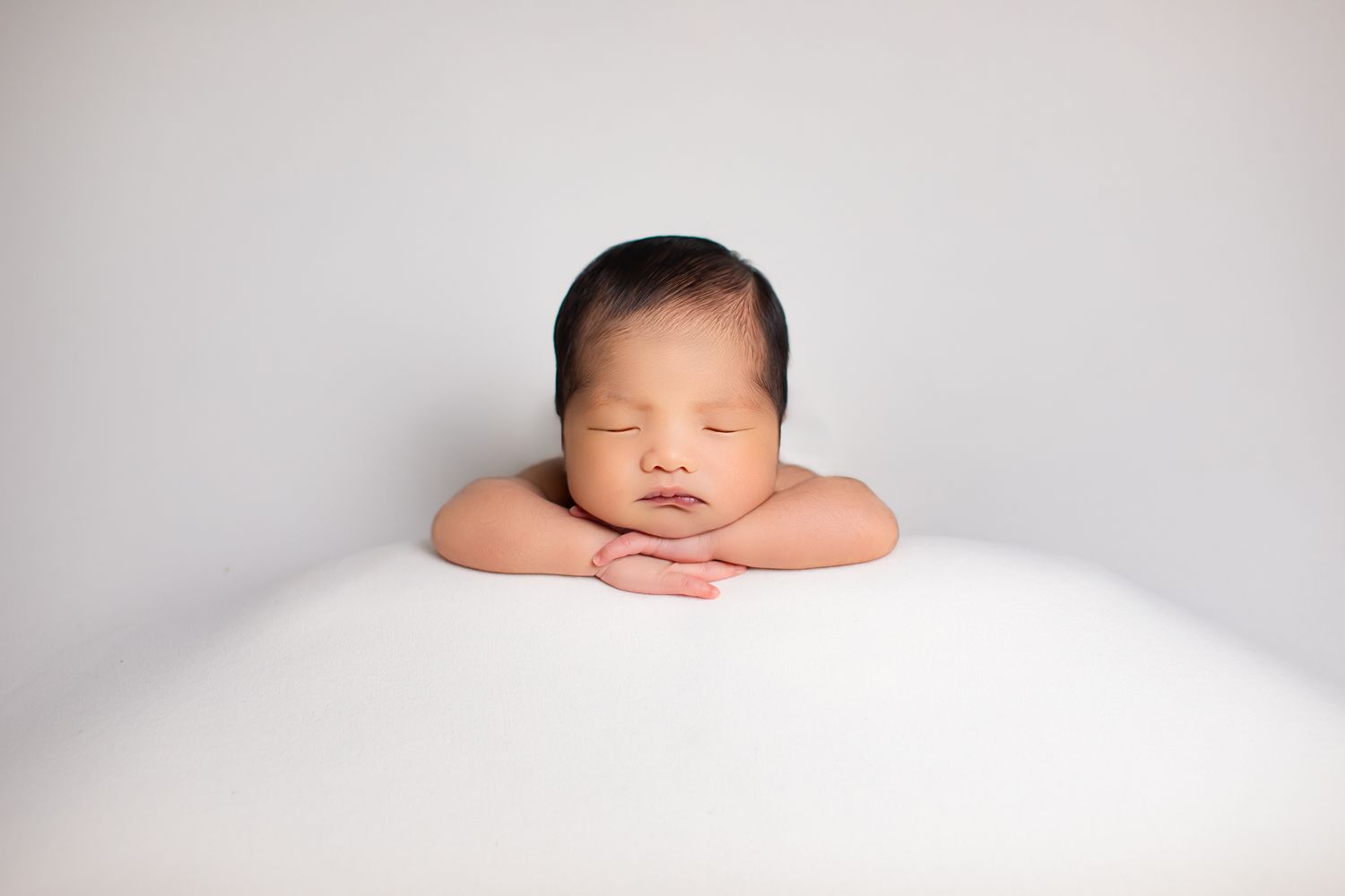 Serene newborn baby resting chin on hands against a clean white background.