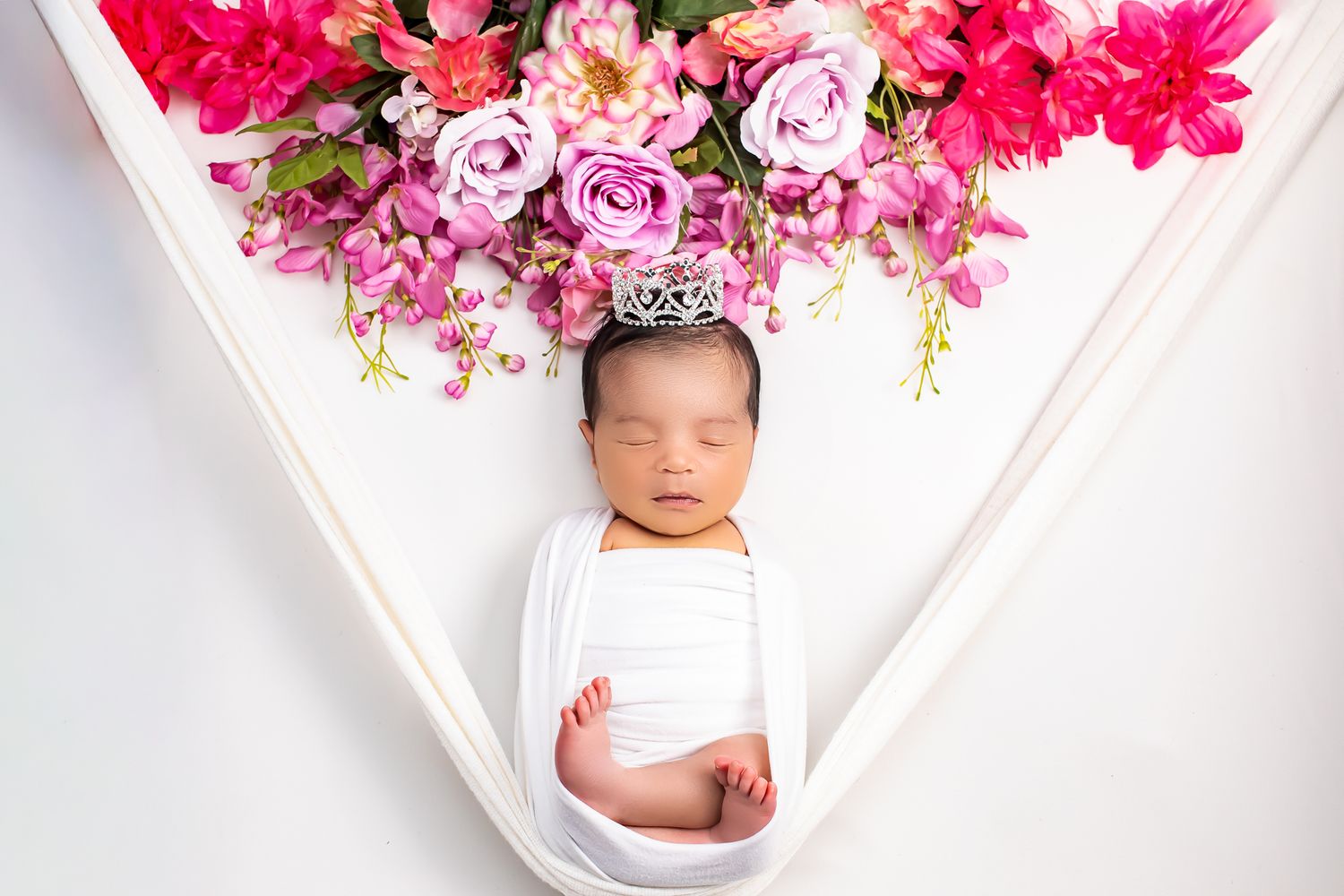 Sleeping newborn baby wrapped in white swaddle wearing tiny crown surrounded by pink and red flowers.