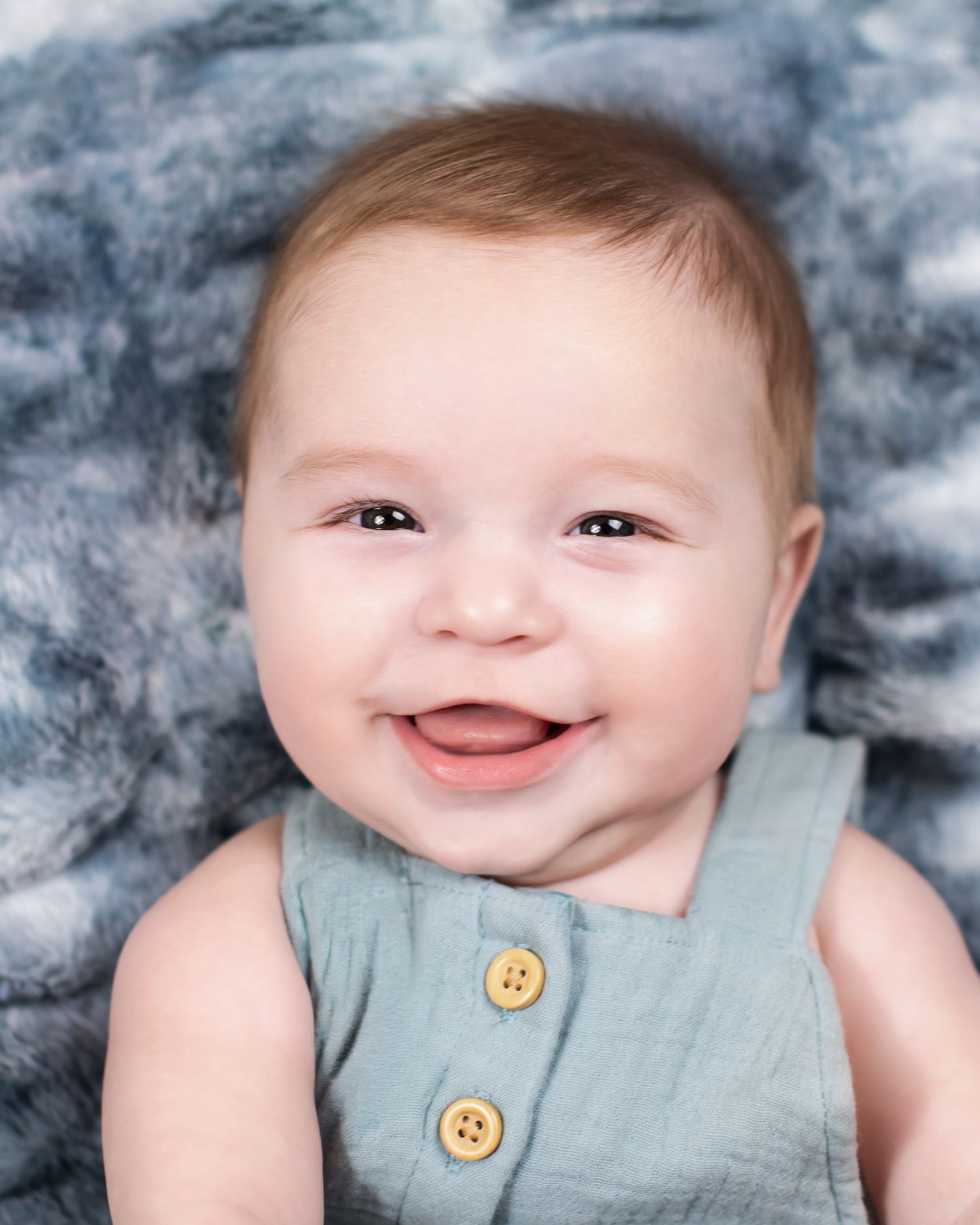 Happy smiling baby in a teal romper with yellow buttons lying on a blue blanket.