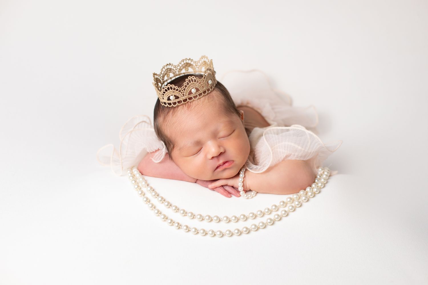 Peaceful newborn baby girl sleeping with gold crown headband and pearl necklace on pink blanket.