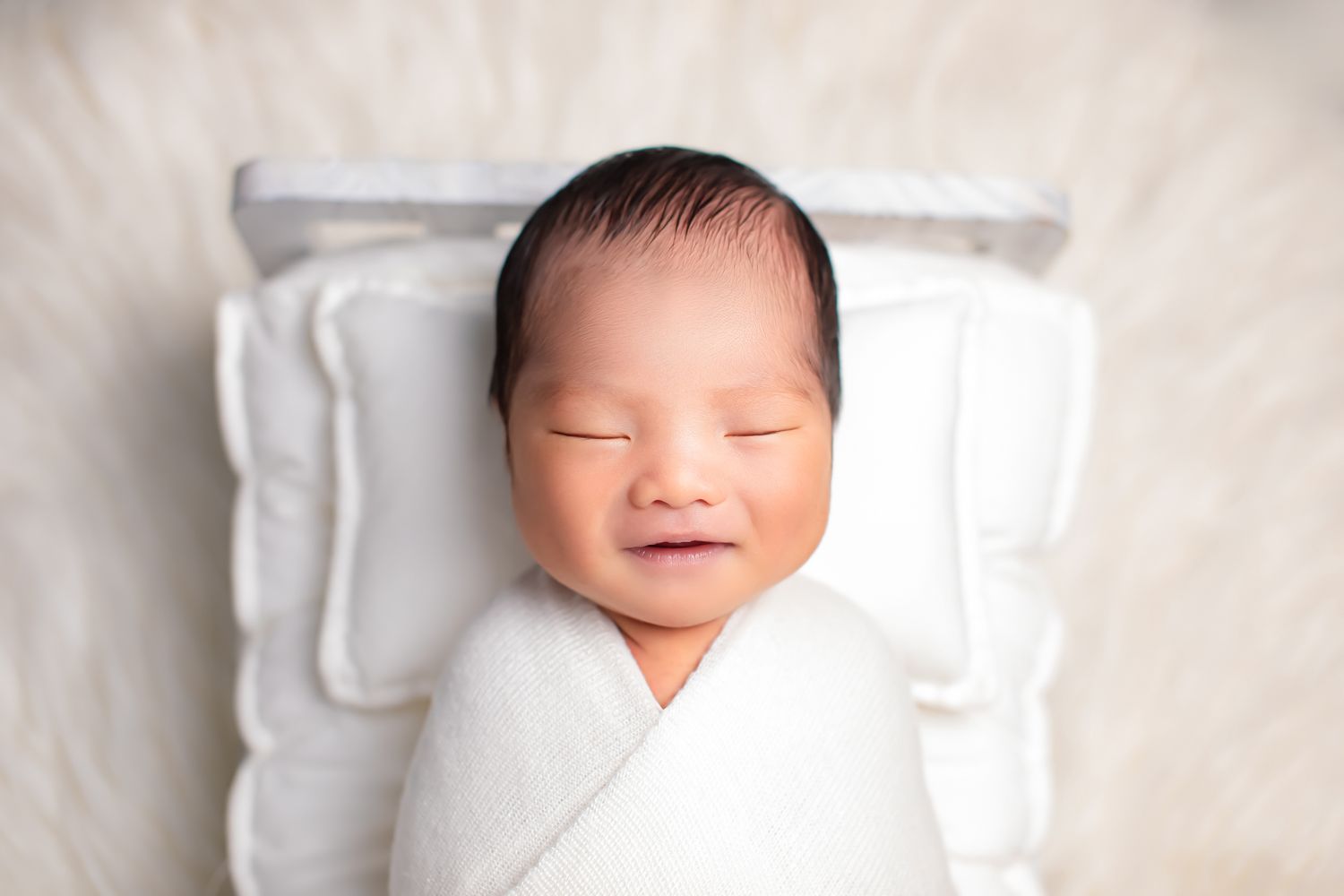 Smiling newborn baby wrapped in white swaddle lying on a tiny white pillow on fluffy white rug.