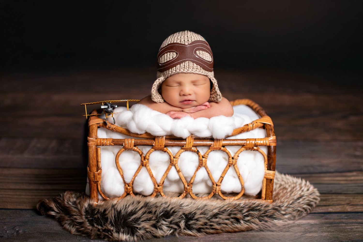 Sleeping newborn baby wearing a knit aviator hat in a rustic wicker basket with white blanket.