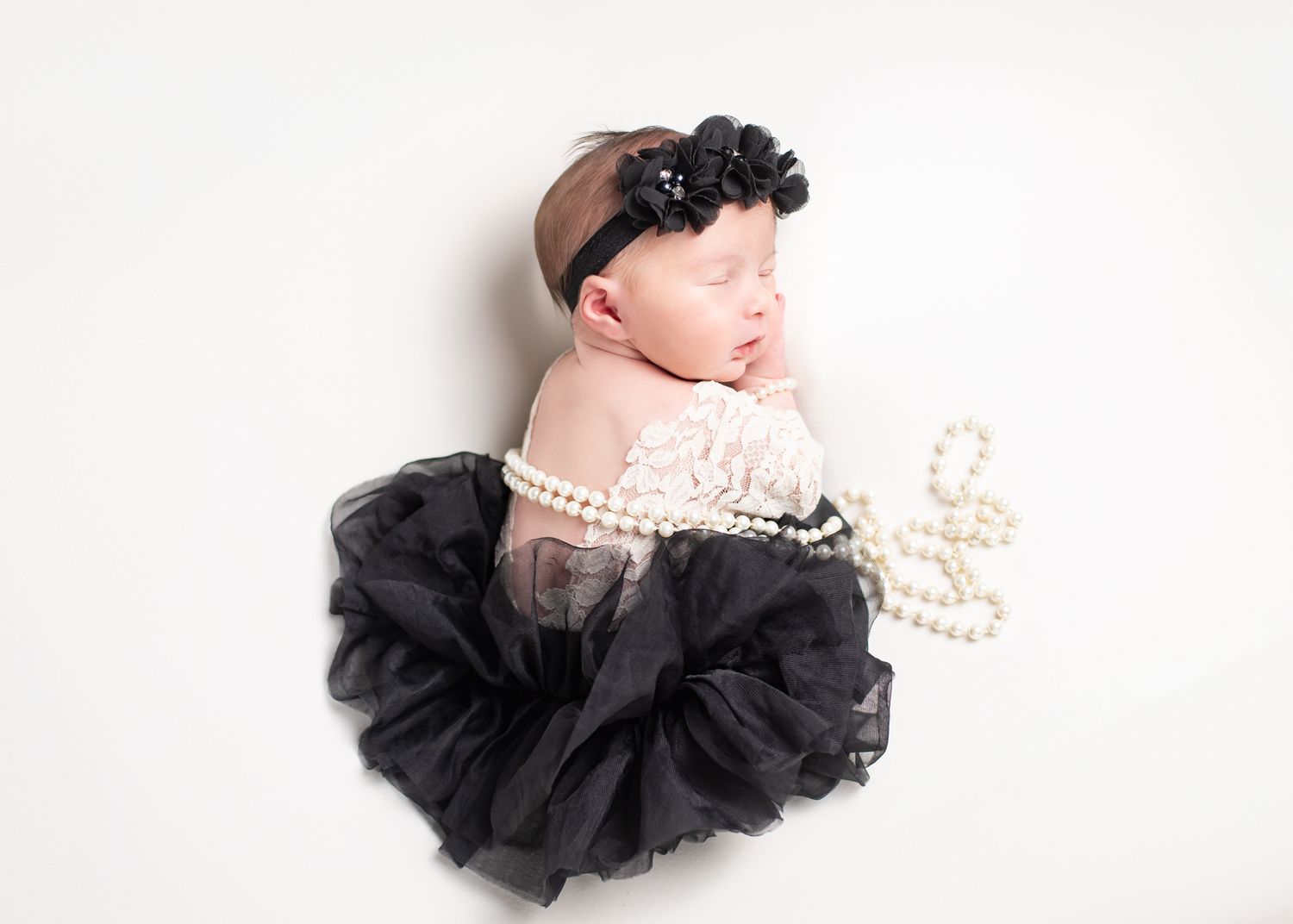 Sleeping newborn baby girl in black tutu dress with pearl necklace and floral headband on white background.