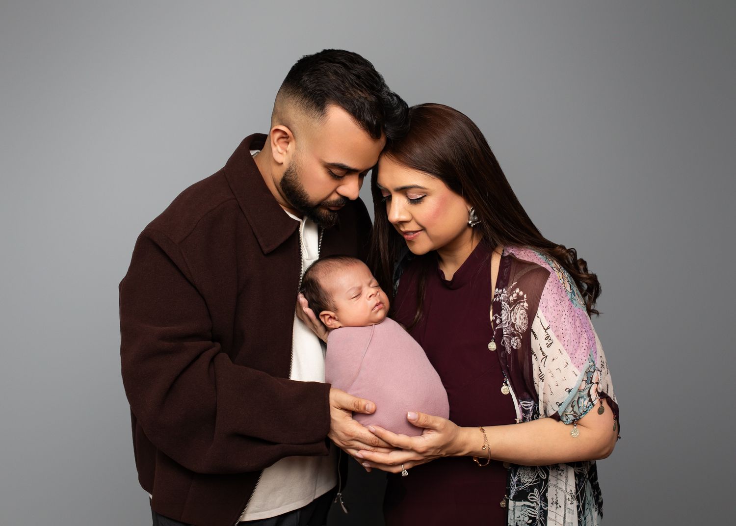 Parents tenderly hold their swaddled newborn baby in a professional studio portrait against a gray background.
