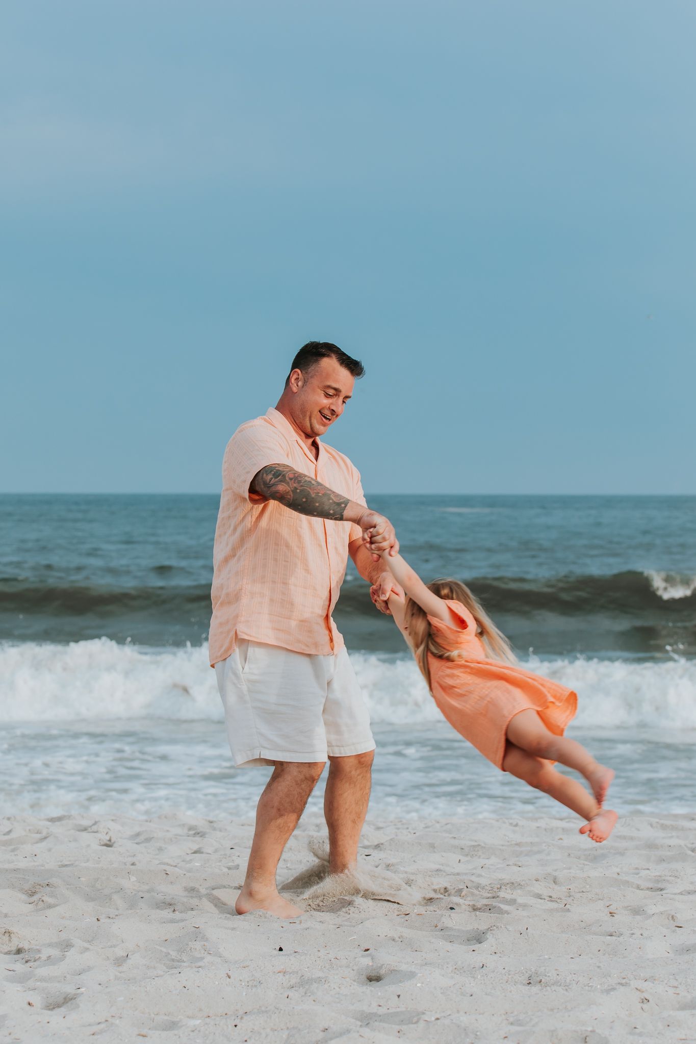 Father joyfully spinning young daughter in orange dress on sandy beach with ocean waves behind them.