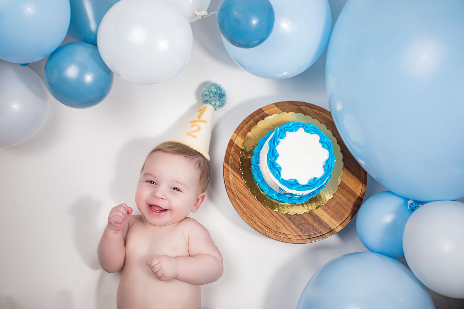 Smiling baby boy in party hat beside blue birthday cake surrounded by blue and white balloons.