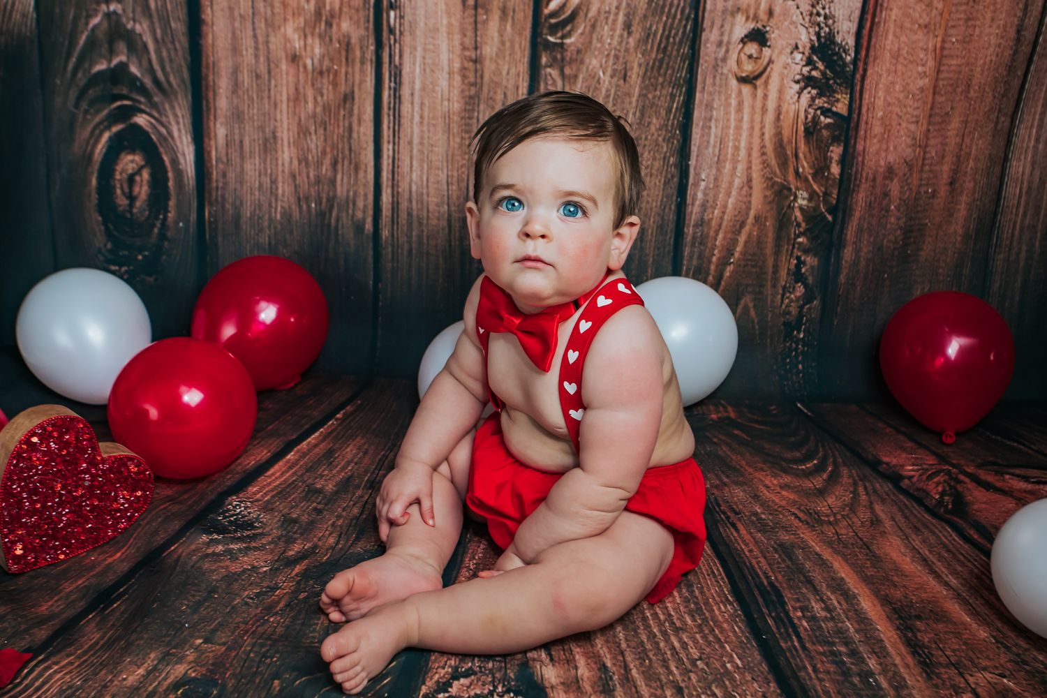 Blue-eyed baby in red outfit sitting on wood floor with red and white balloons for Valentine's Day photo.