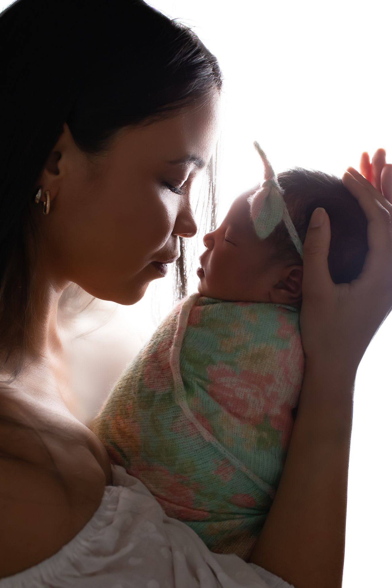 Mother tenderly holds her swaddled newborn baby close to her face in a softly backlit portrait.