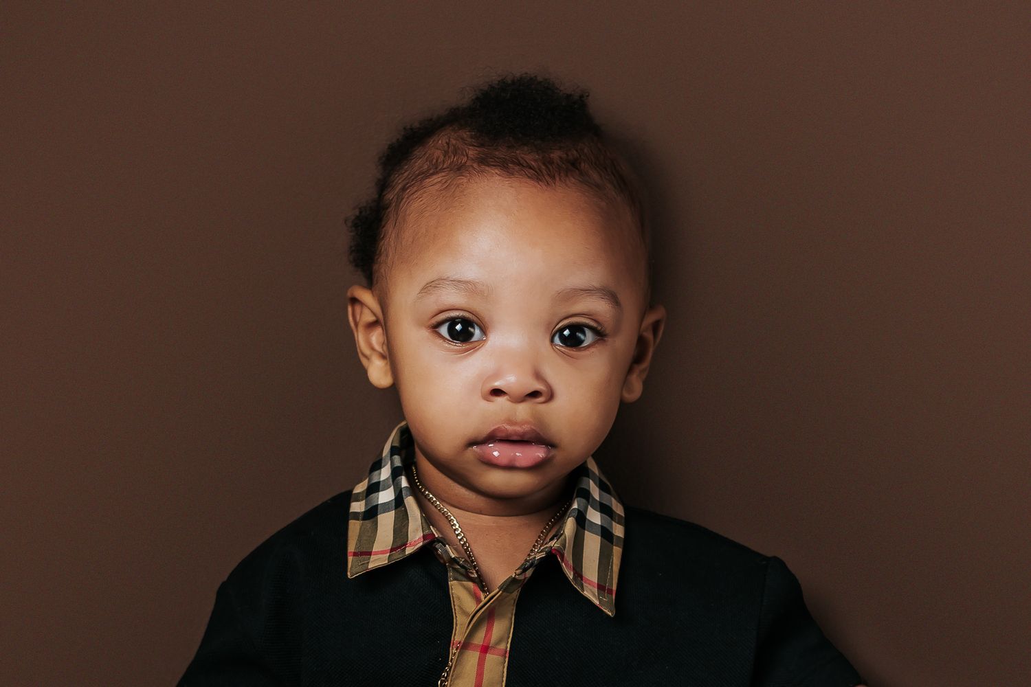 Adorable baby boy in a Burberry collar black shirt posing against a warm brown background.