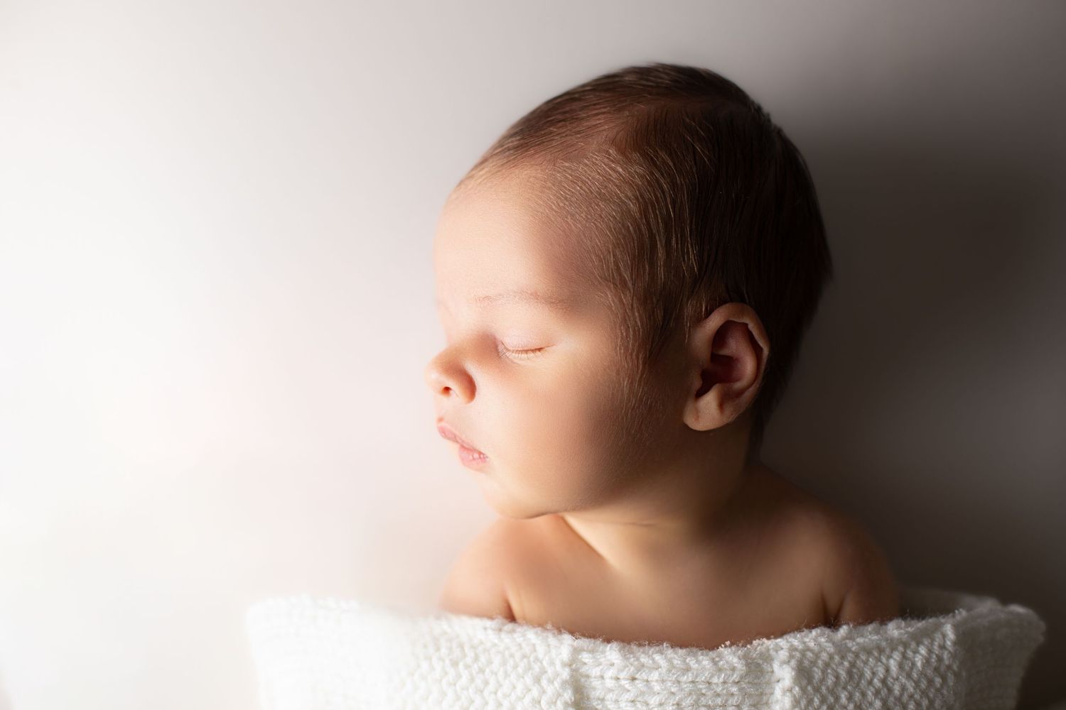 Peaceful newborn baby sleeping wrapped in a soft white knit blanket against a light background.