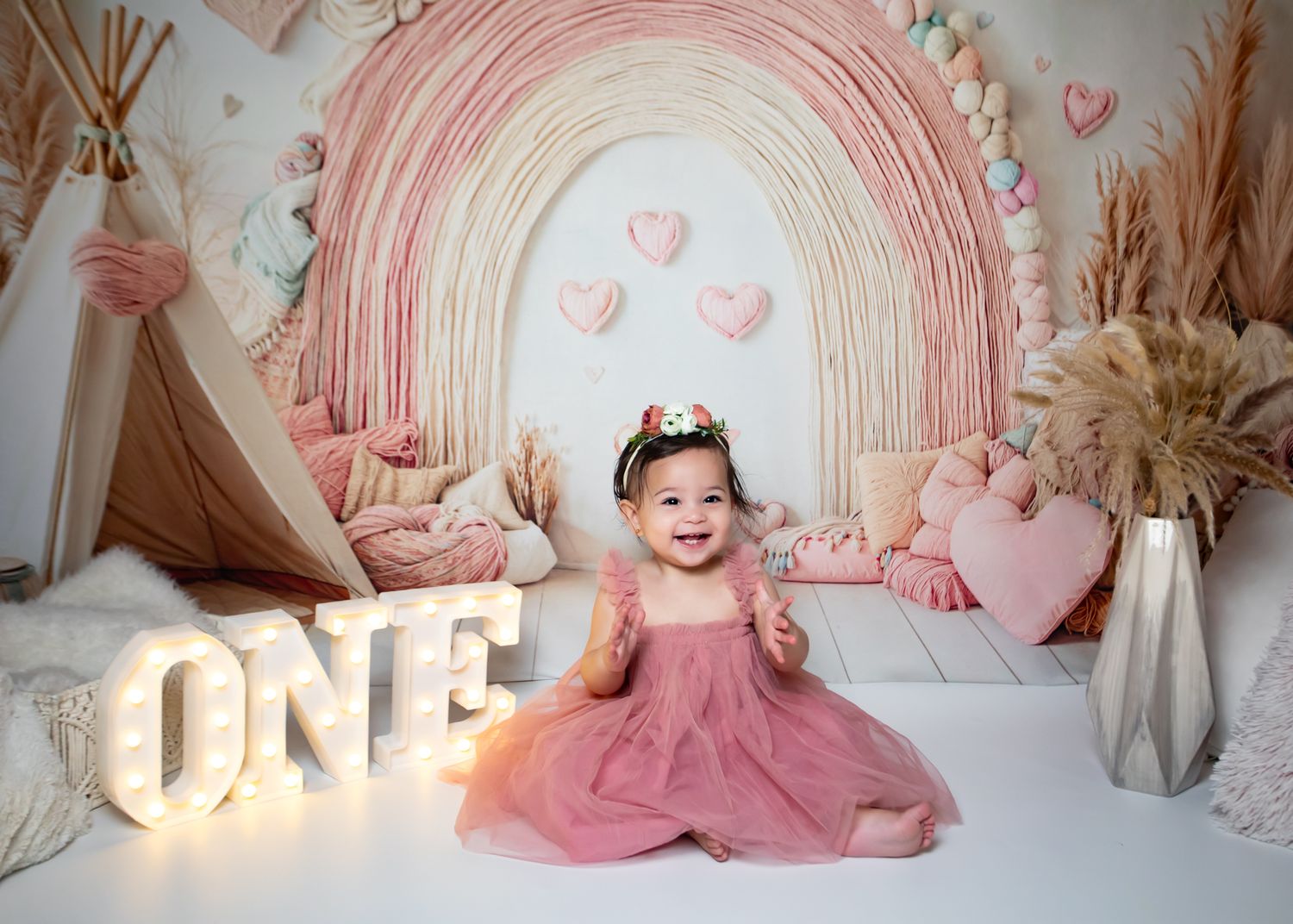 Smiling baby girl in pink dress celebrating first birthday with ONE sign and rainbow boho backdrop.