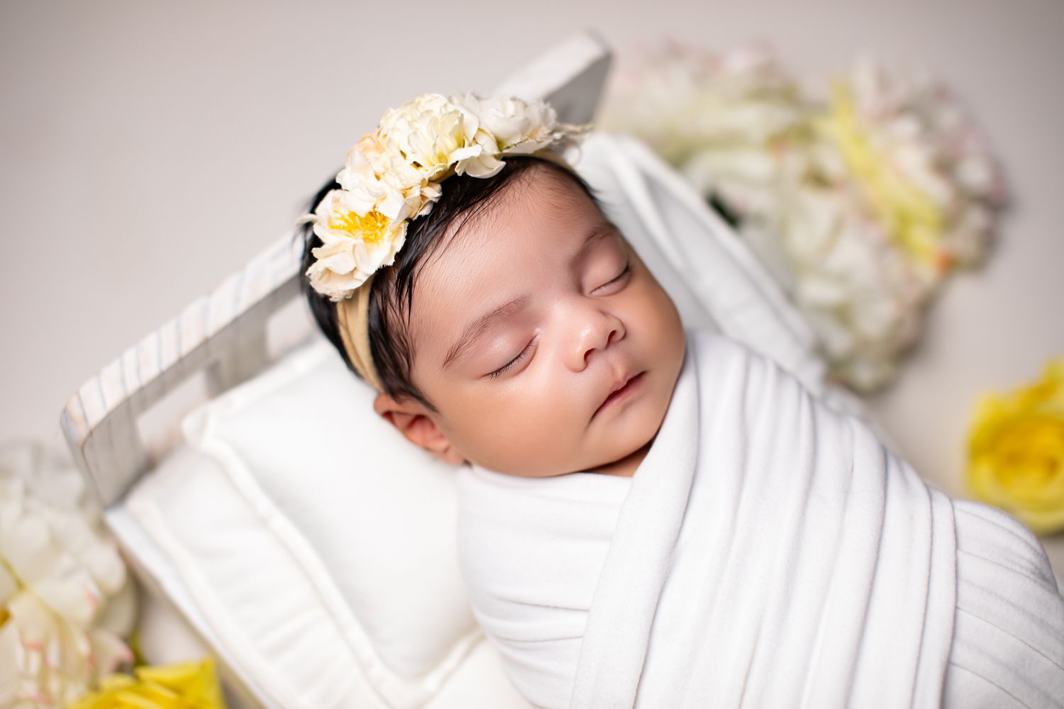 Peaceful newborn baby wrapped in white swaddle wearing a yellow floral headband in a tiny bed.