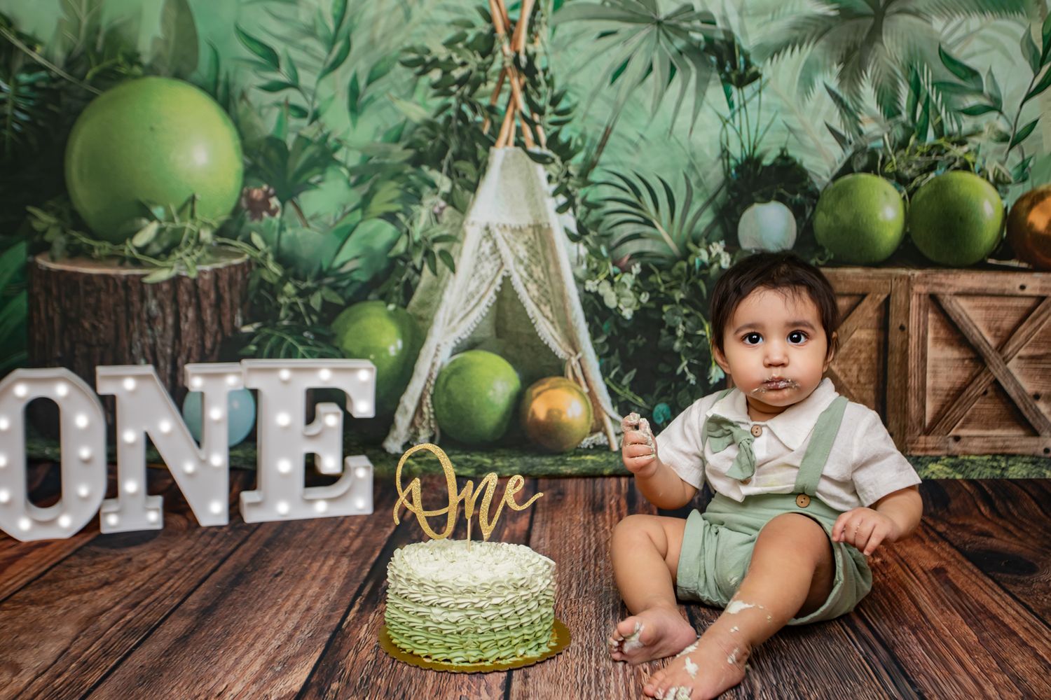 Baby boy in green outfit at ONE-themed jungle birthday cake smash with teepee backdrop.