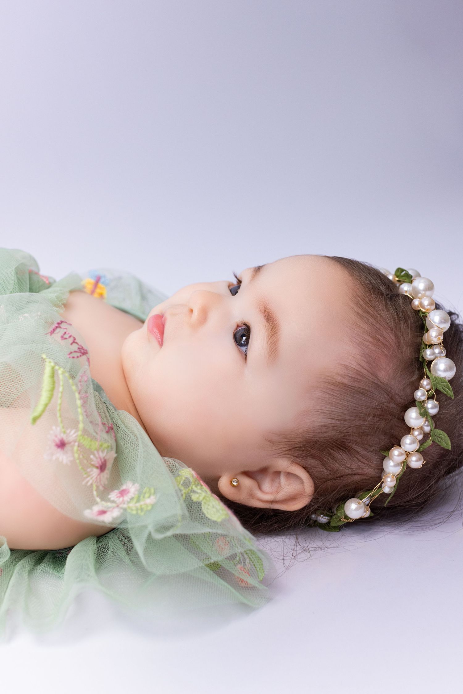 Adorable baby girl in green floral dress and pearl headband lying down, looking up with big brown eyes.
