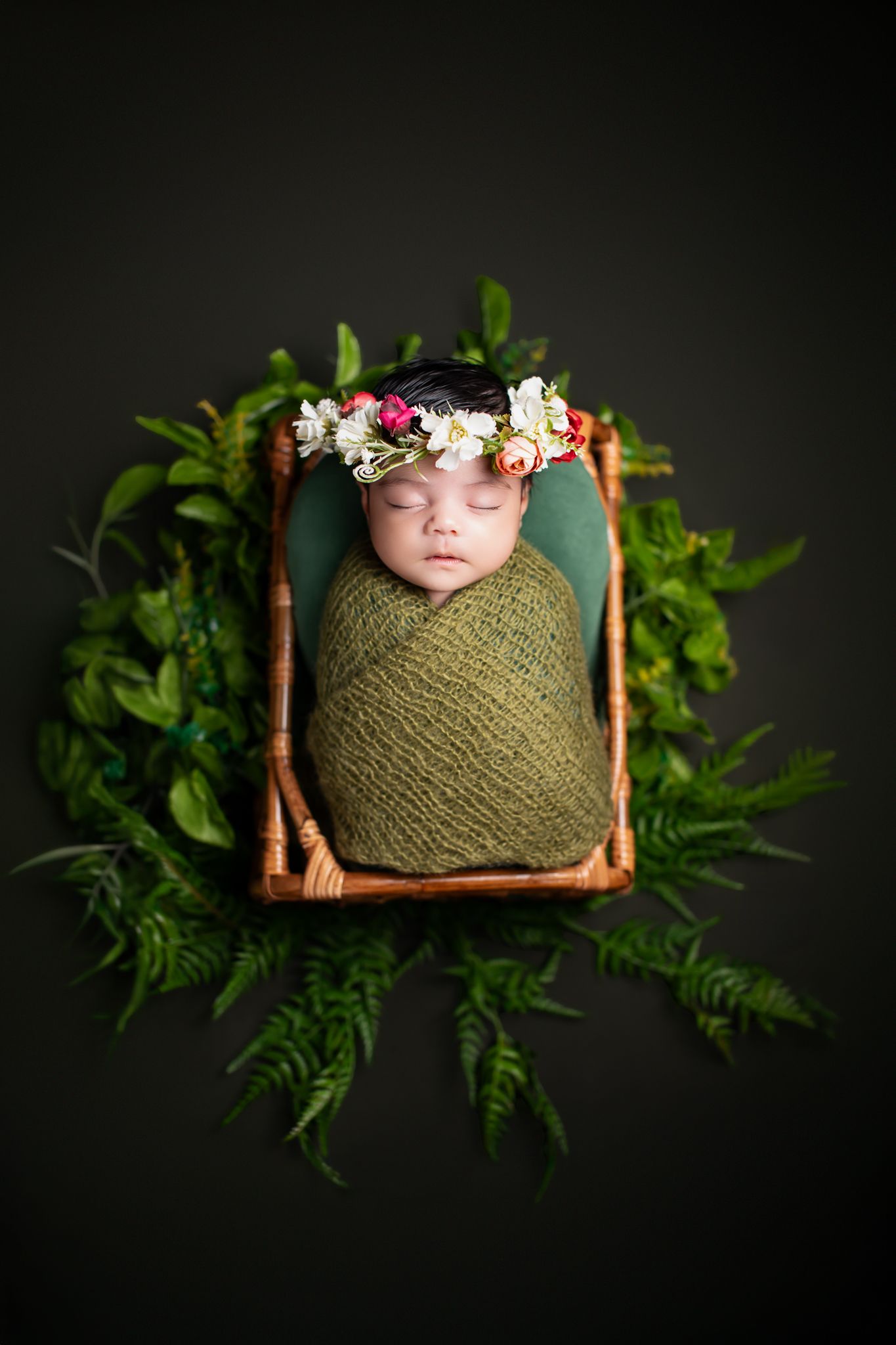 Sleeping newborn swaddled in green wrap with floral crown in a basket surrounded by ferns.