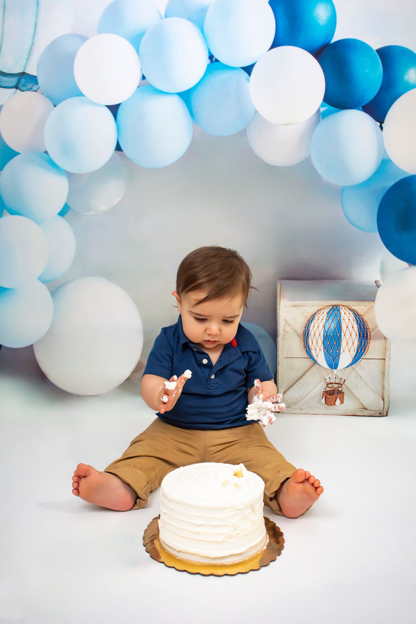 Baby boy sitting with smash cake at first birthday surrounded by blue and white balloons.