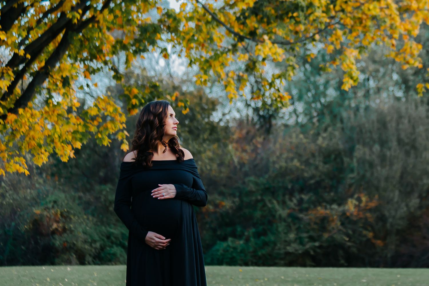 Pregnant woman in black off-shoulder dress stands in autumn park cradling her baby bump.