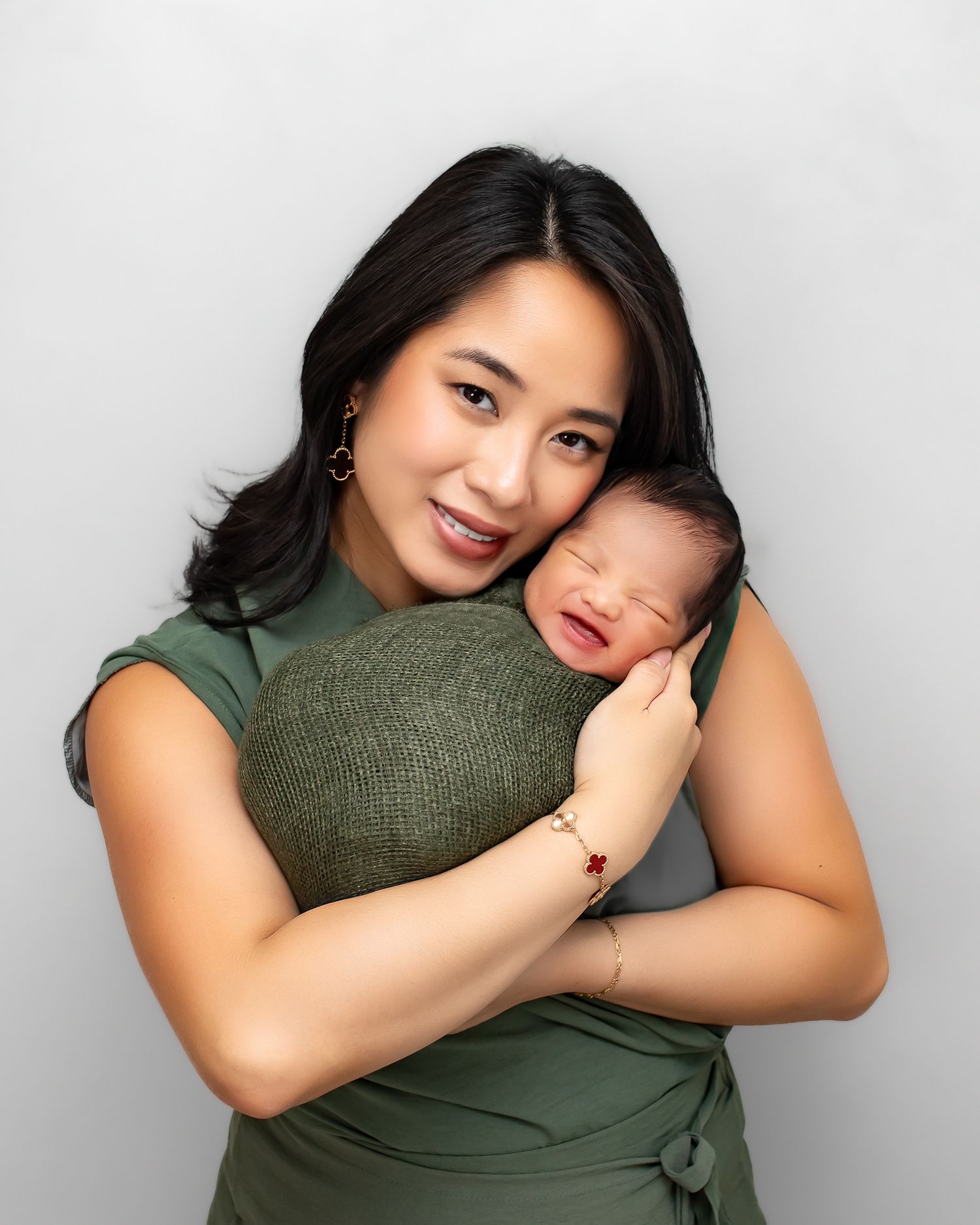 Happy Asian mother in green dress smiles while holding her crying swaddled newborn baby close.
