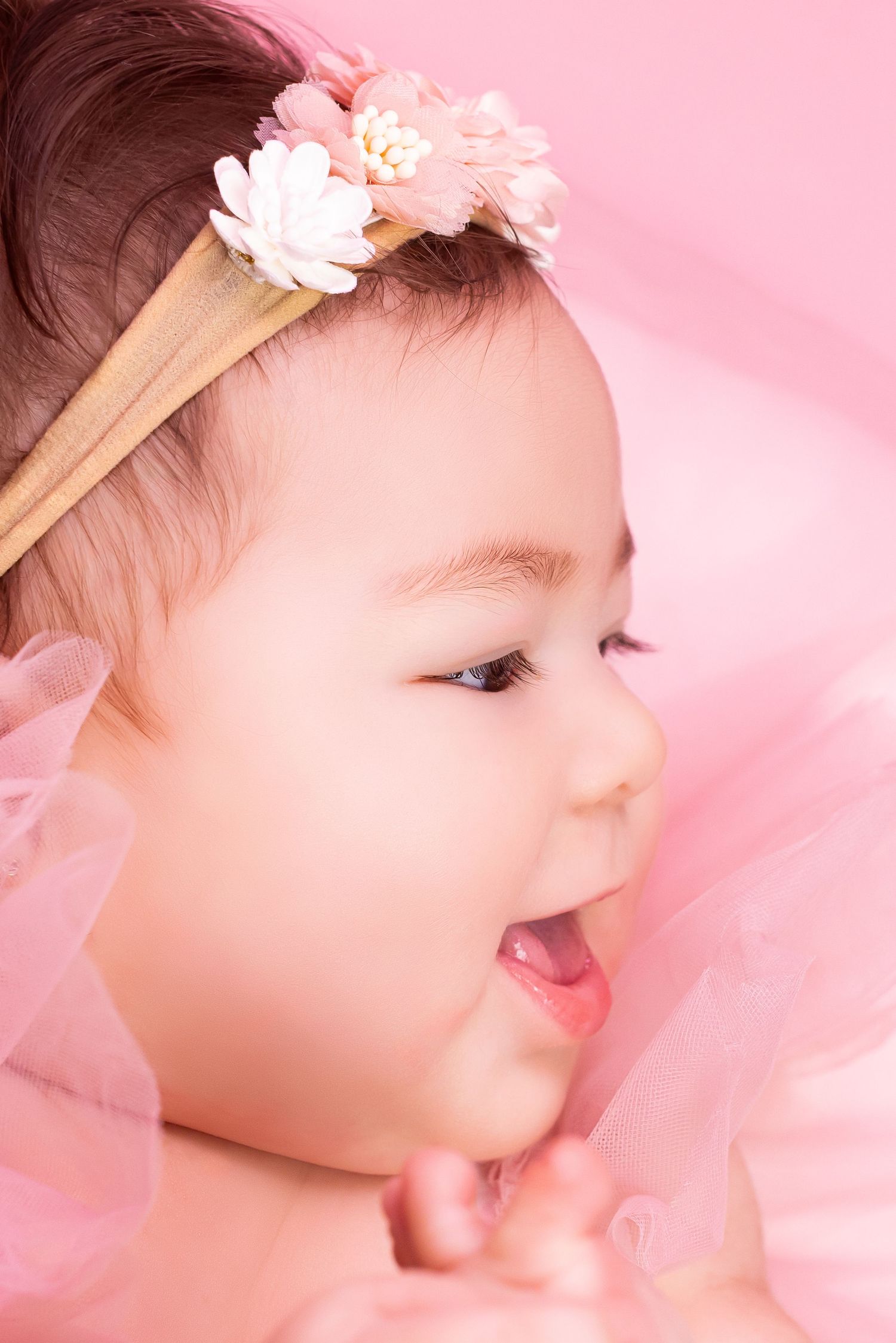 Adorable baby girl wearing a floral headband, smiling joyfully against a soft pink background.