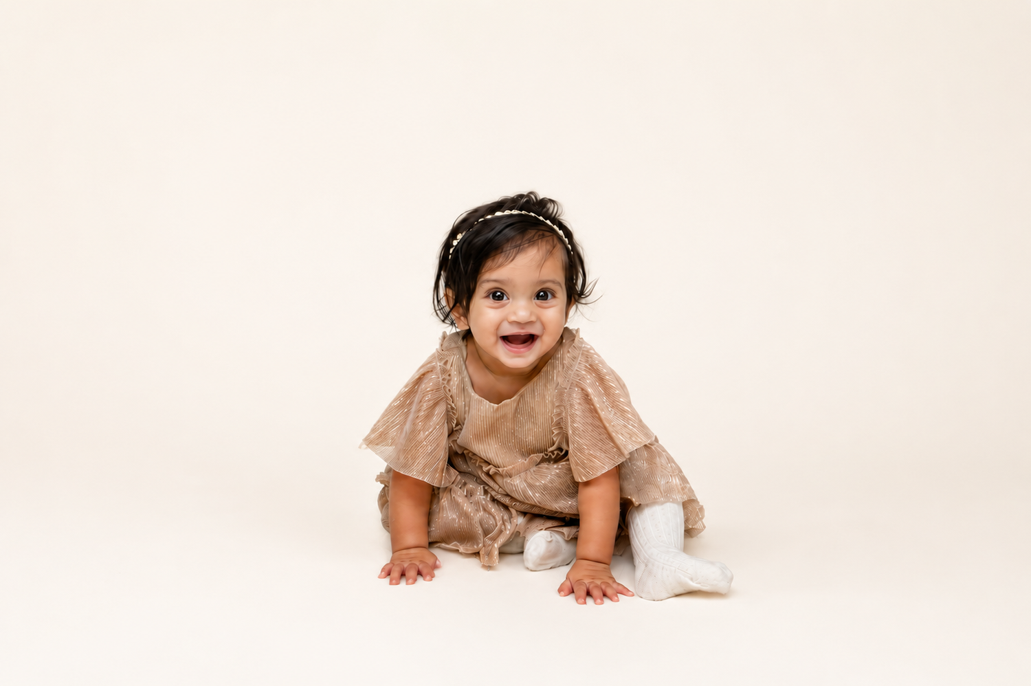 Happy baby girl in brown dress and headband crawling on cream background in professional photo.