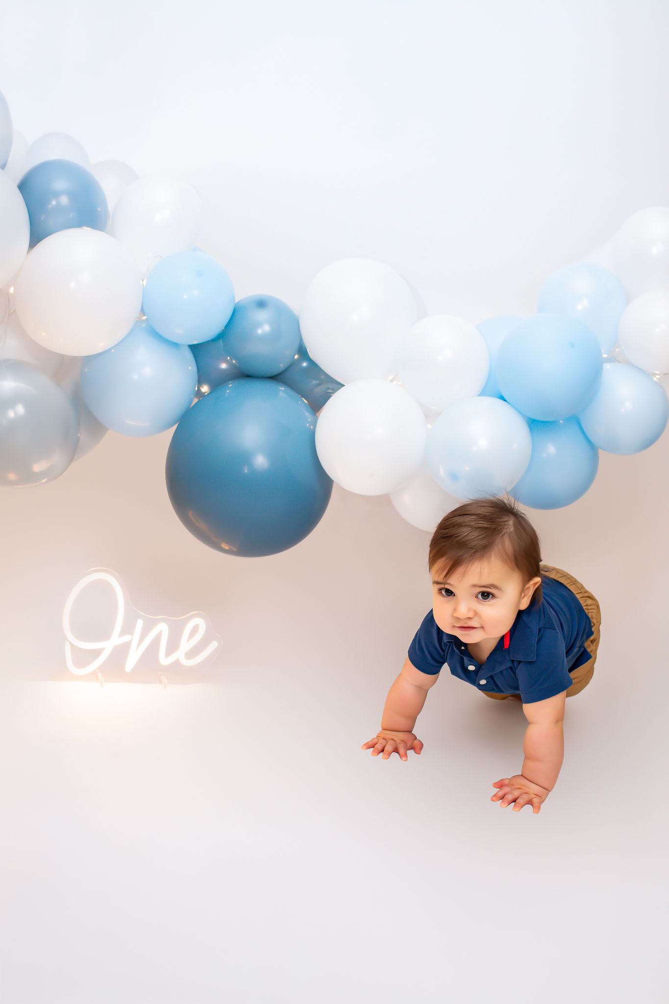 Baby boy at first birthday crawling near blue and white balloon garland with neon One sign.