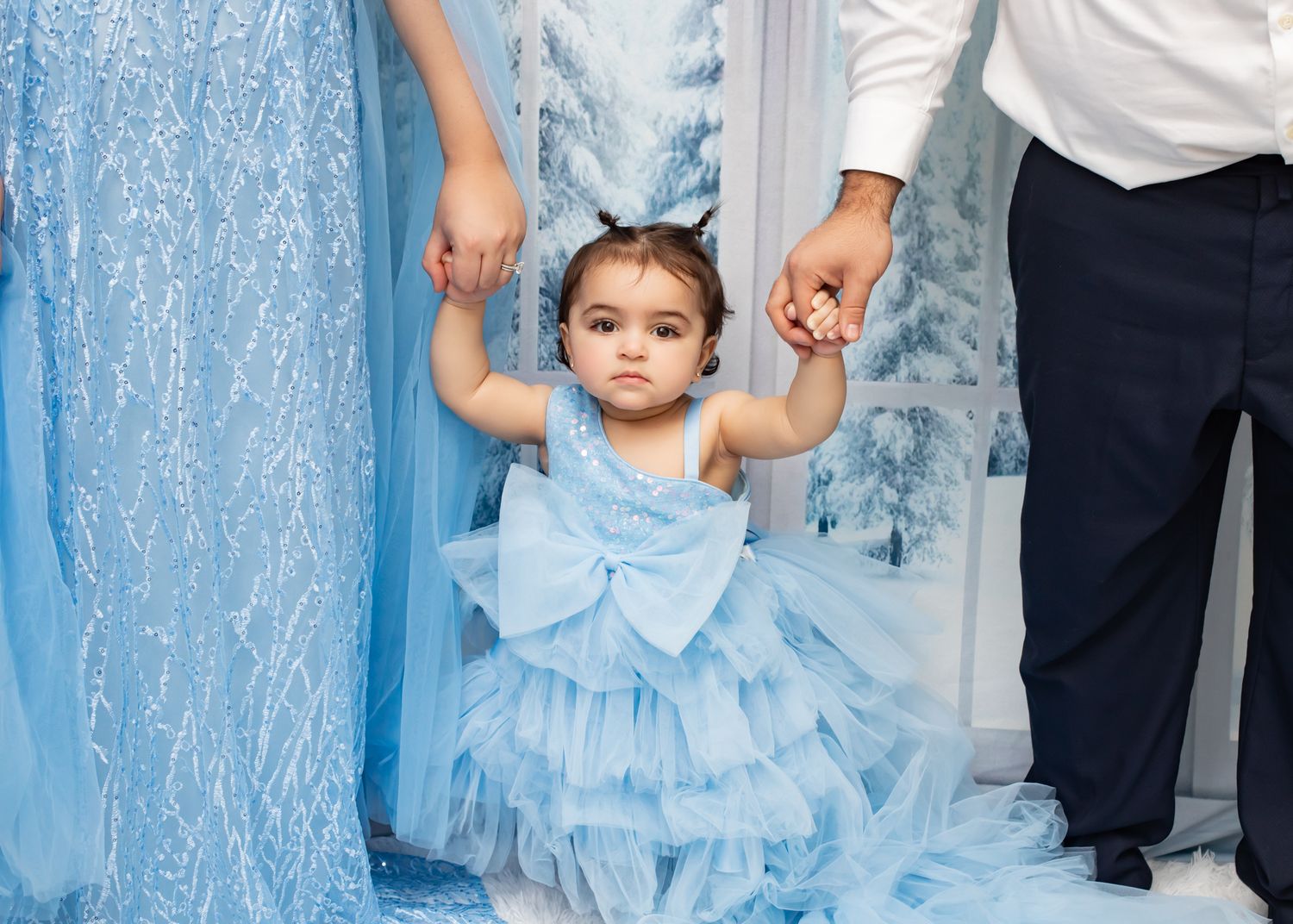 Cute toddler girl in blue tulle dress holding parents hands in a winter wonderland studio setting.