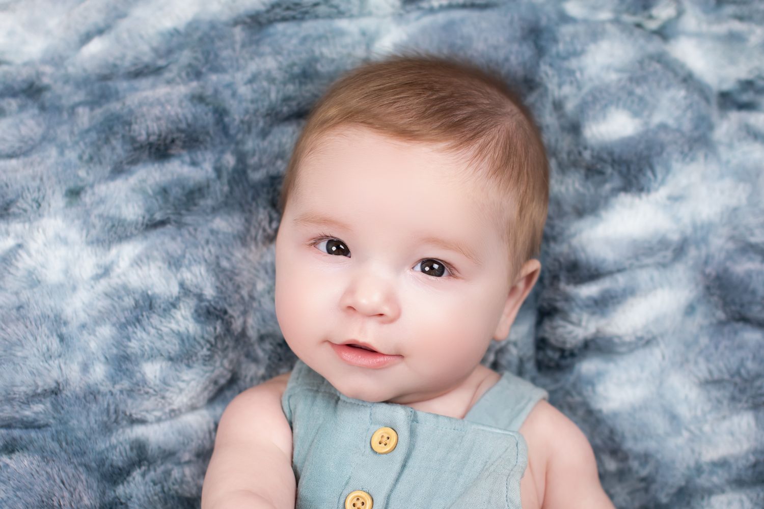 Adorable baby in a teal romper lying on a soft blue fur blanket, looking up with bright curious eyes.