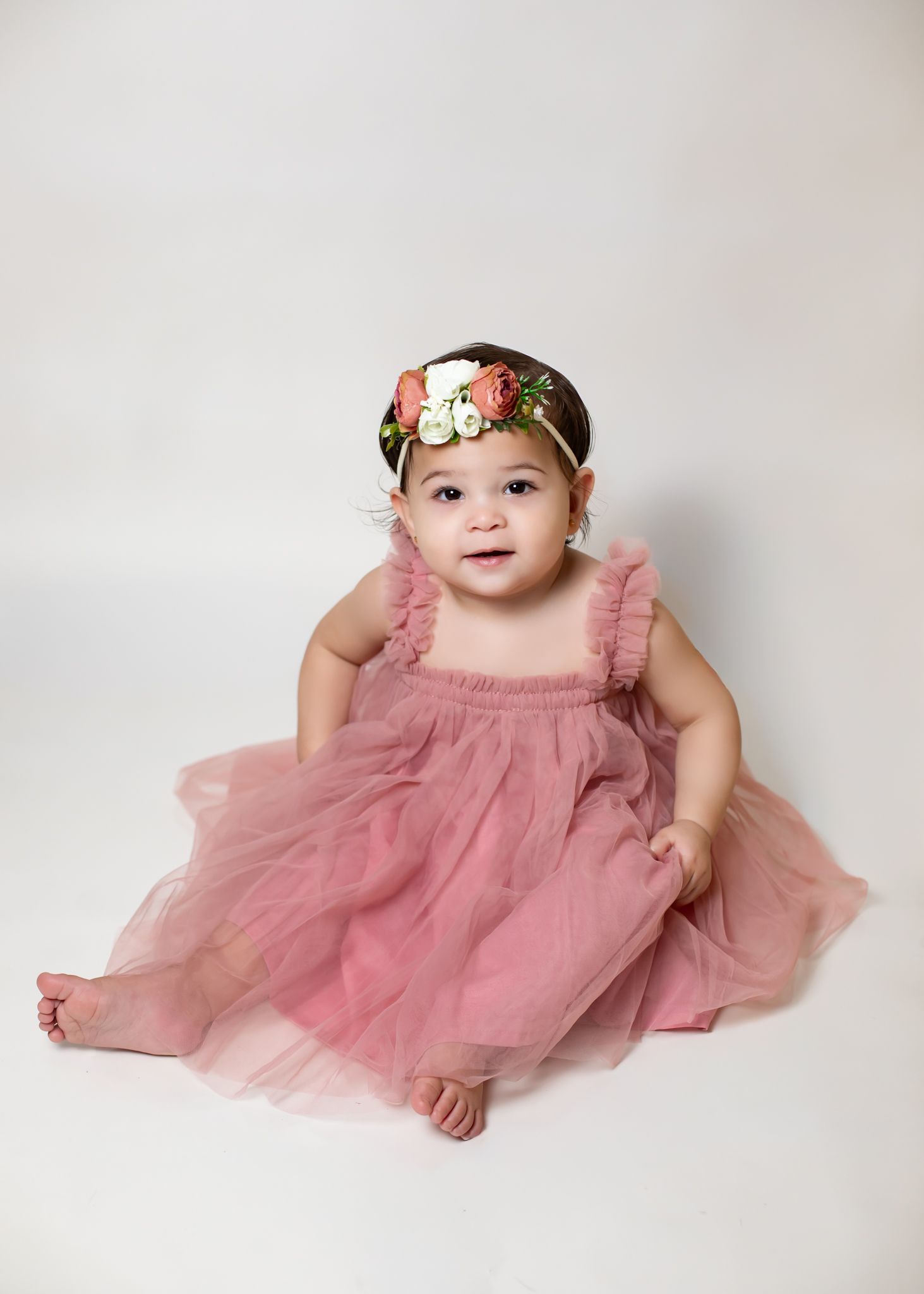 Sweet baby girl in a pink tulle dress and floral headband sitting on a white studio backdrop.